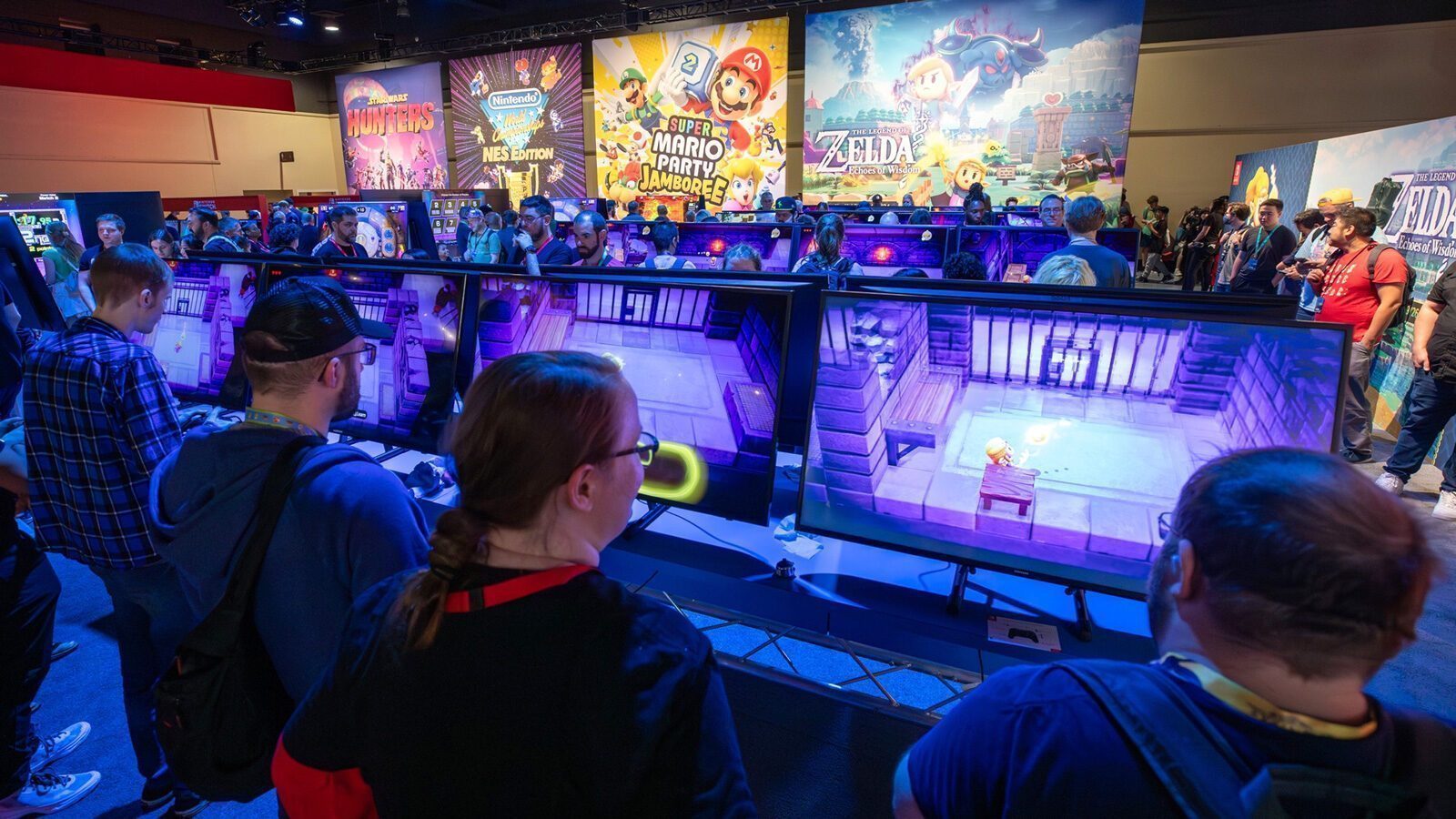 At this must-list event on 08-28-2025, people play video games at a convention with large screens and promotional posters for Nintendo games like Super Mario Party and The Legend of Zelda in the background.