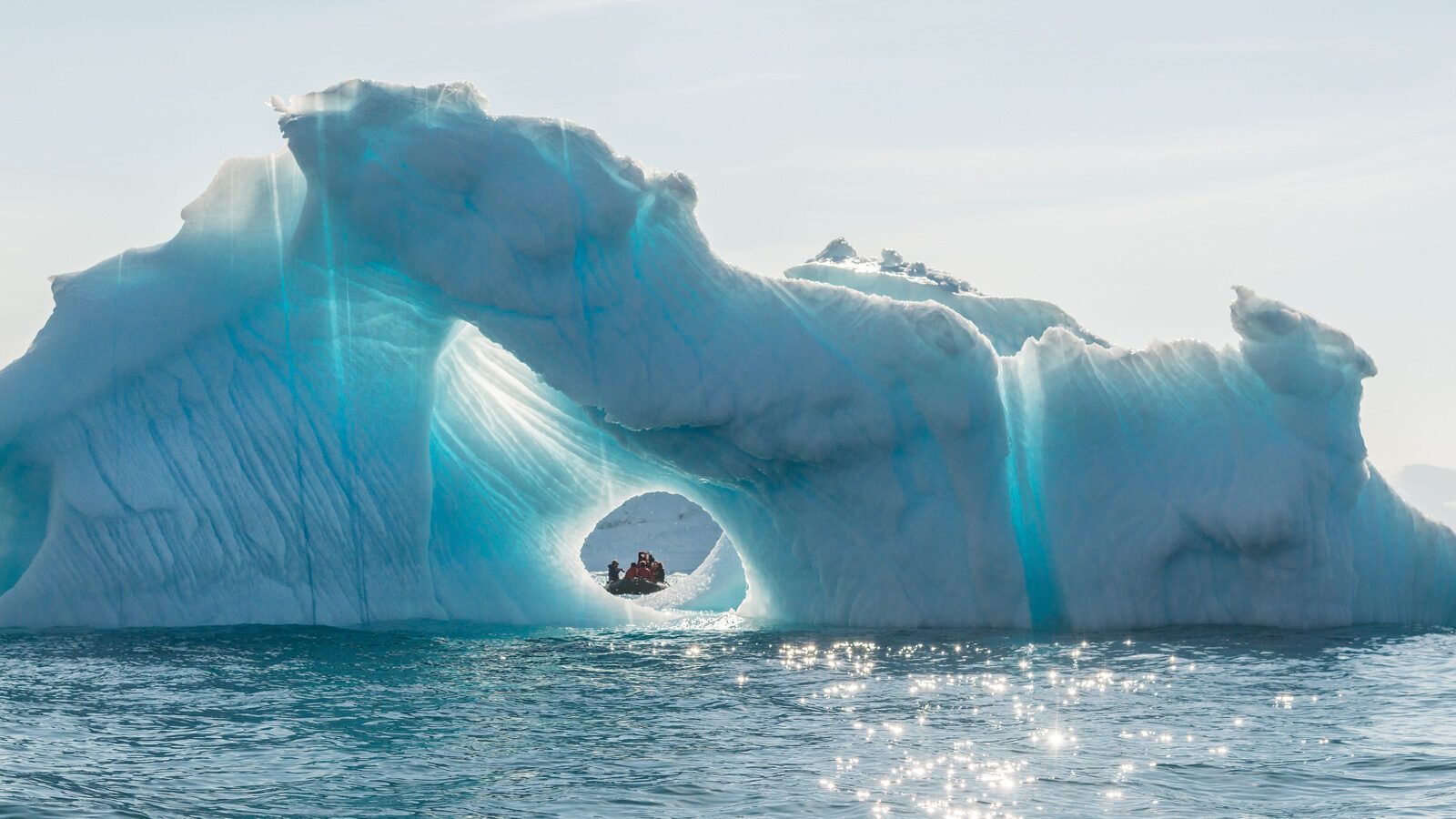 A small boat with people is visible through a round hole in a large floating iceberg, surrounded by calm ocean water—one of the unforgettable sights awaiting travelers on United flights to Greenland.
