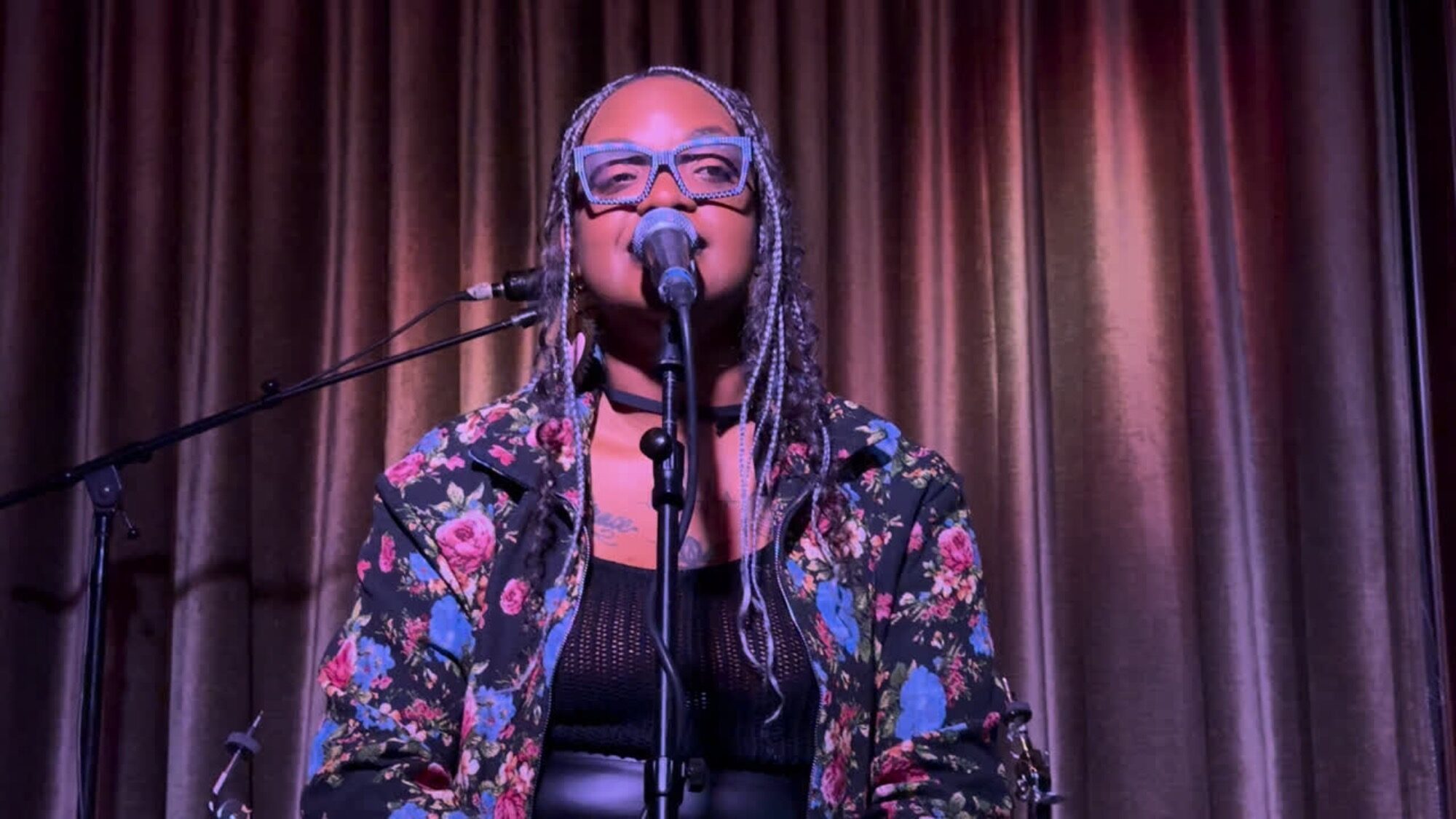 A person with braided hair and blue glasses sings into a microphone on stage, wearing a floral jacket and black top, with a curtain backdrop.