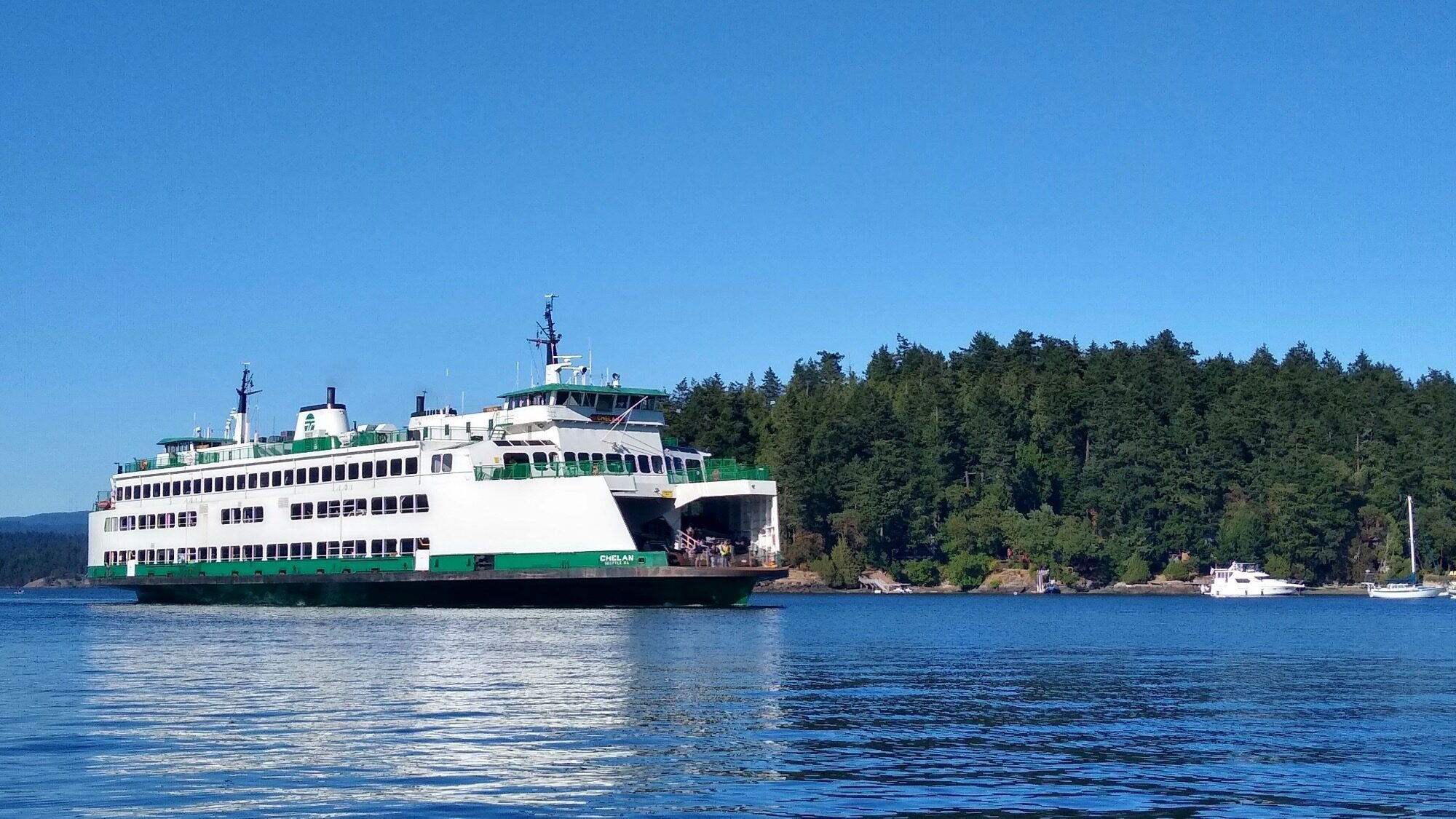 All Aboard! A large white and green ferry travels on calm water near a forested shoreline under a clear blue sky.