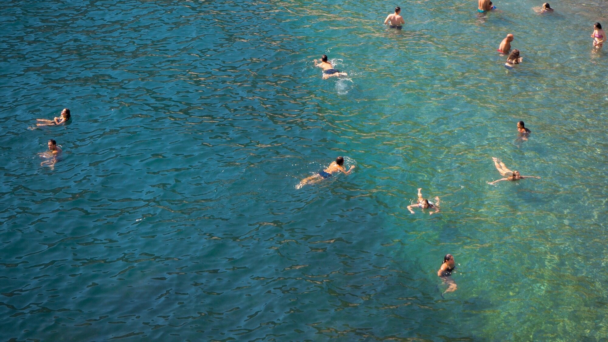 A group of people swim and float in clear blue-green water, viewed from above.