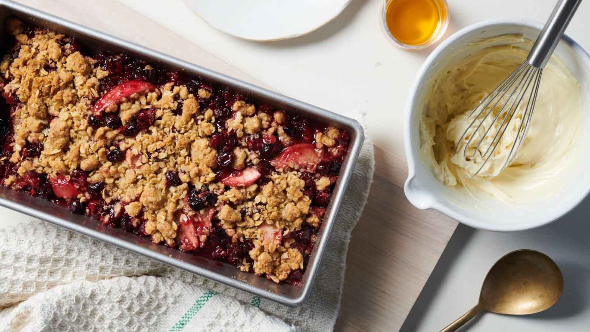 A rectangular baking dish filled with fruit crumble sits next to a bowl of whipped cream, a whisk, a spoon, and a small cup of syrup—perfect for enjoying during berry season.