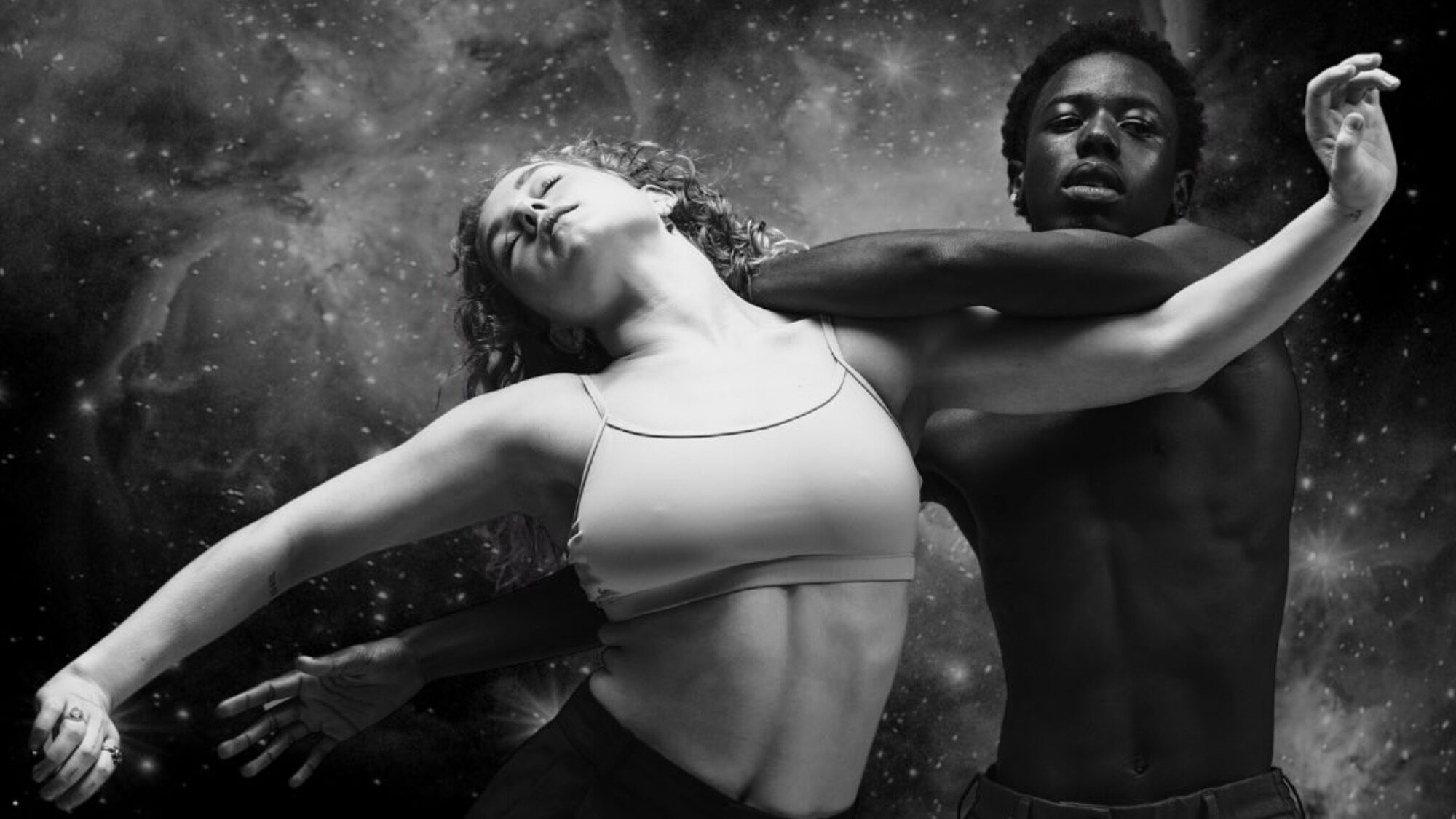 Two dancers, one male and one female, perform an expressive pose against a cosmic, star-filled background in black and white.
