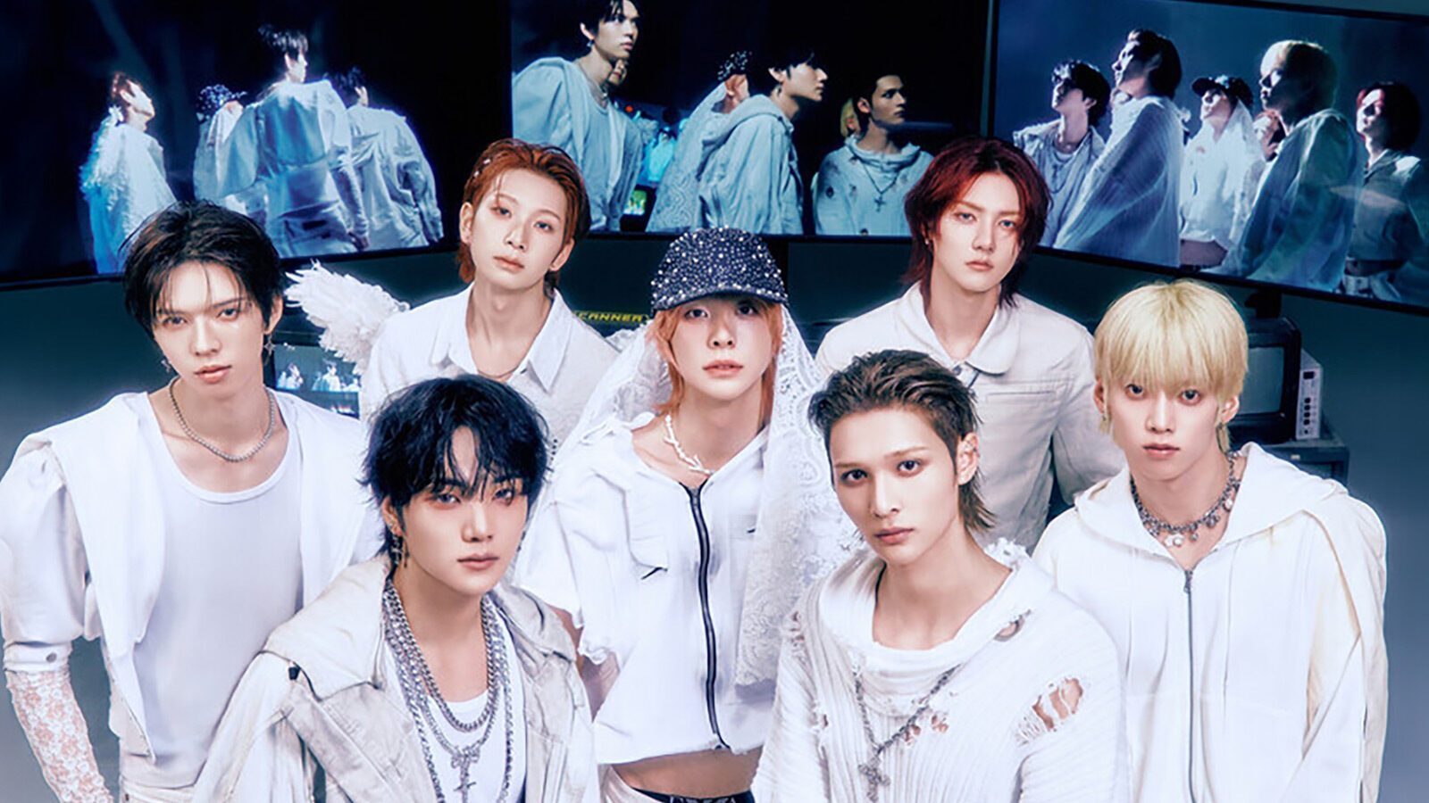 Seven young men dressed in white outfits pose for a group photo indoors, with screens in the background displaying their image.