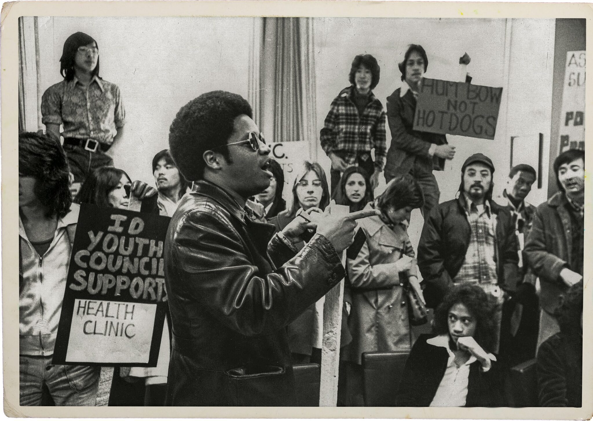 A group of people, some holding protest signs, gather indoors; one man in sunglasses and a leather jacket speaks about resilience while others listen or hold signs reflecting their resistance.