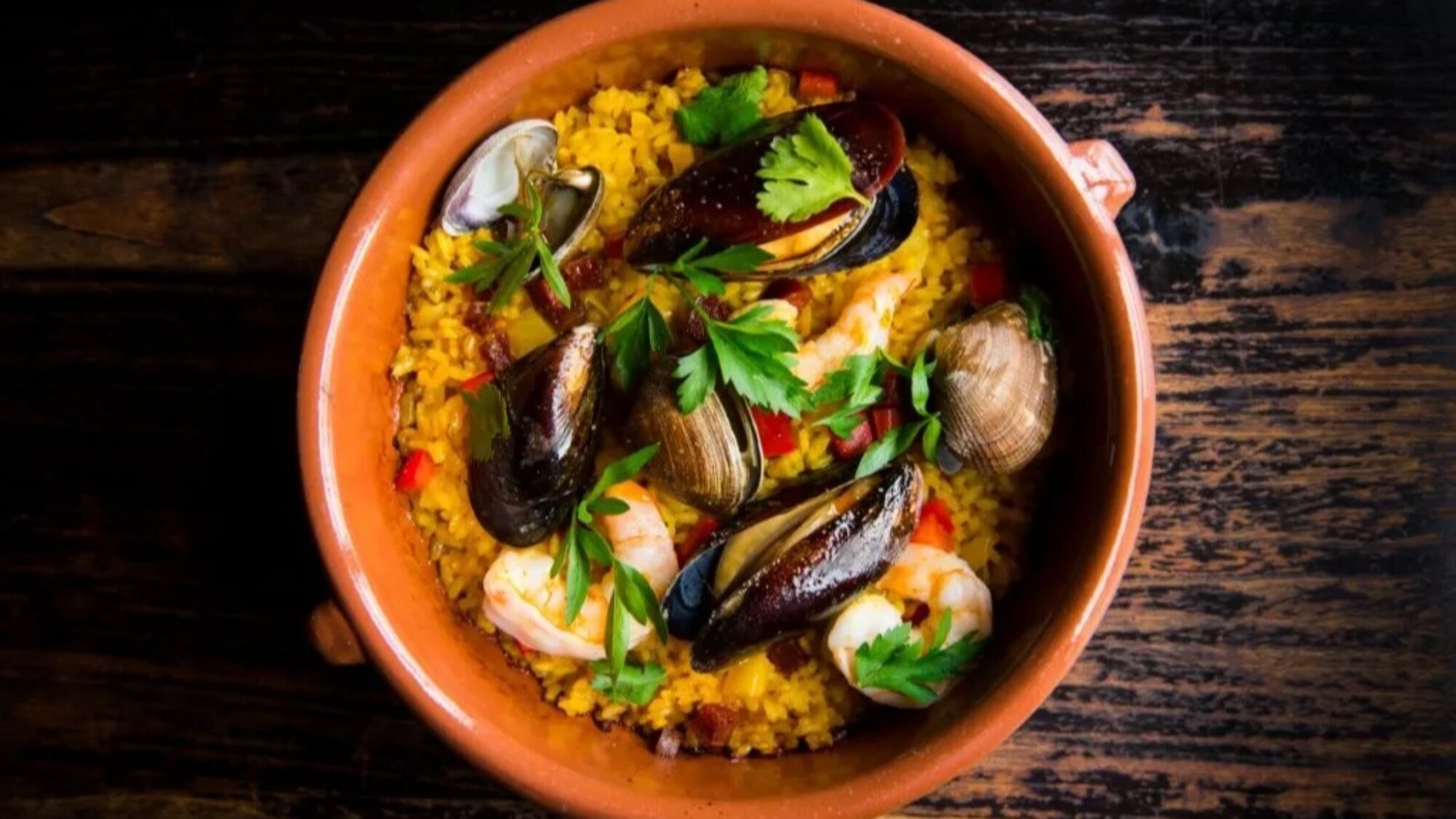 A brown ceramic bowl with seafood paella, featuring rice, mussels, clams, shrimp, red peppers, and parsley, set on a dark wooden surface.