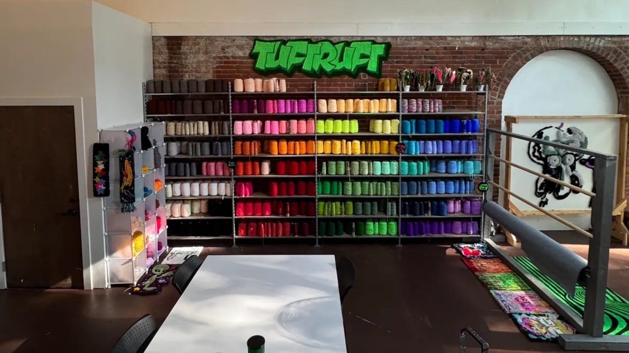 Shelves filled with yarn in a gradient of colors, a tufting gun rack, and a white table in a studio labeled "TUFT" with a green sign on an exposed brick wall—Tuft Stuff is one of the creative things to do in Seattle.