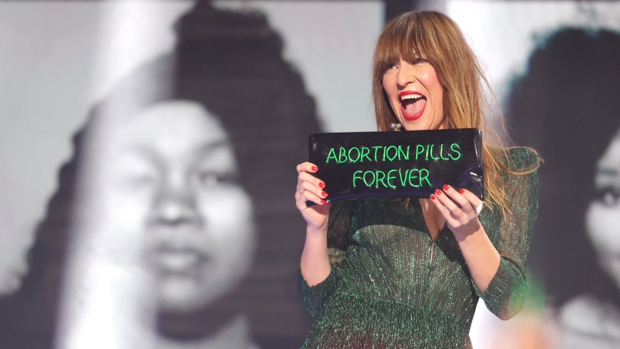 A woman in a green dress holds a black sign with green writing that reads "ABORTION PILLS FOREVER," standing in front of black-and-white portrait images.