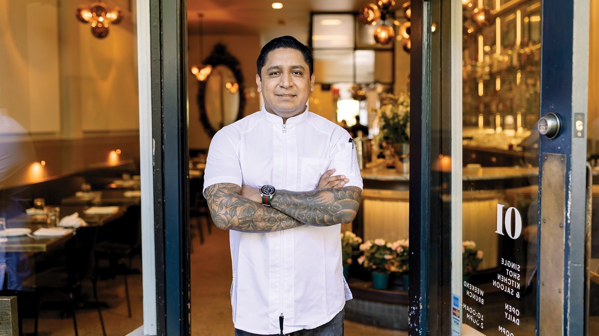 A man with tattooed arms, exuding endurance, stands at the restaurant entrance in a white short-sleeve chef's jacket, smiling with arms crossed—a true masterclass in culinary confidence.