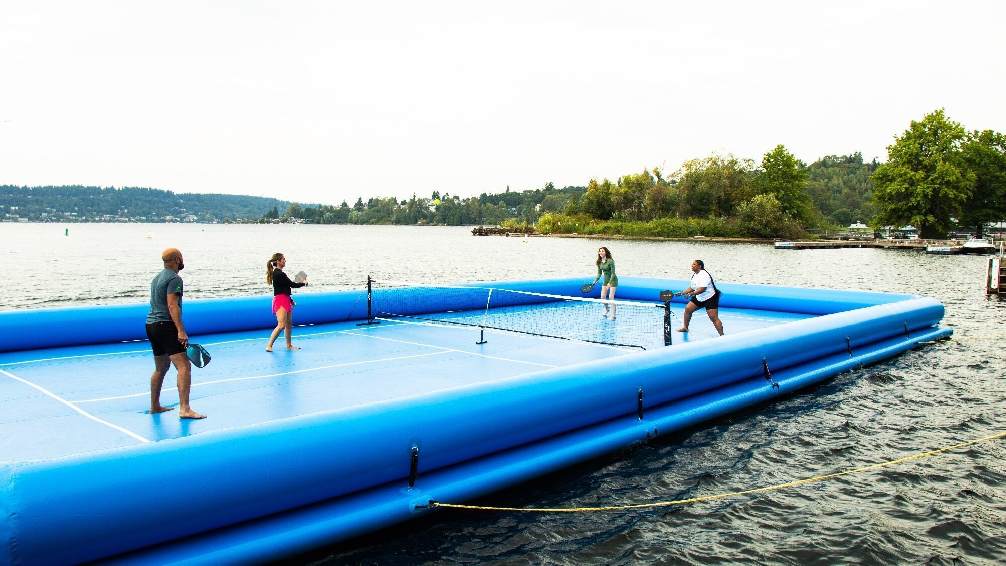 Four people play pickleball on a blue inflatable court floating on a lake, with trees and a shoreline visible in the background.