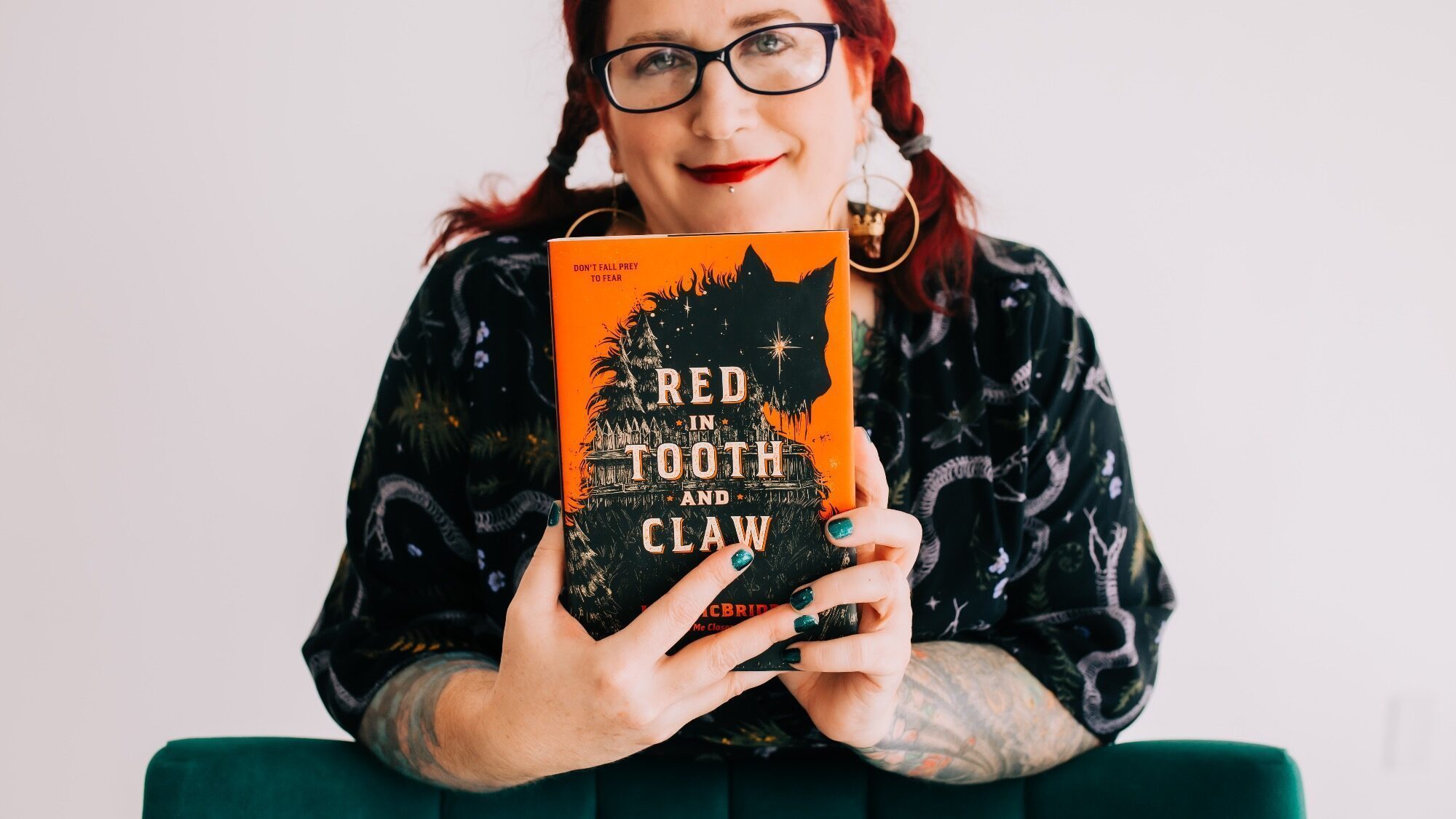 A person with glasses and red hair holds up "Red in Tooth and Claw" by author Lish McBride, a fiction book featuring a black animal silhouette on its orange cover.