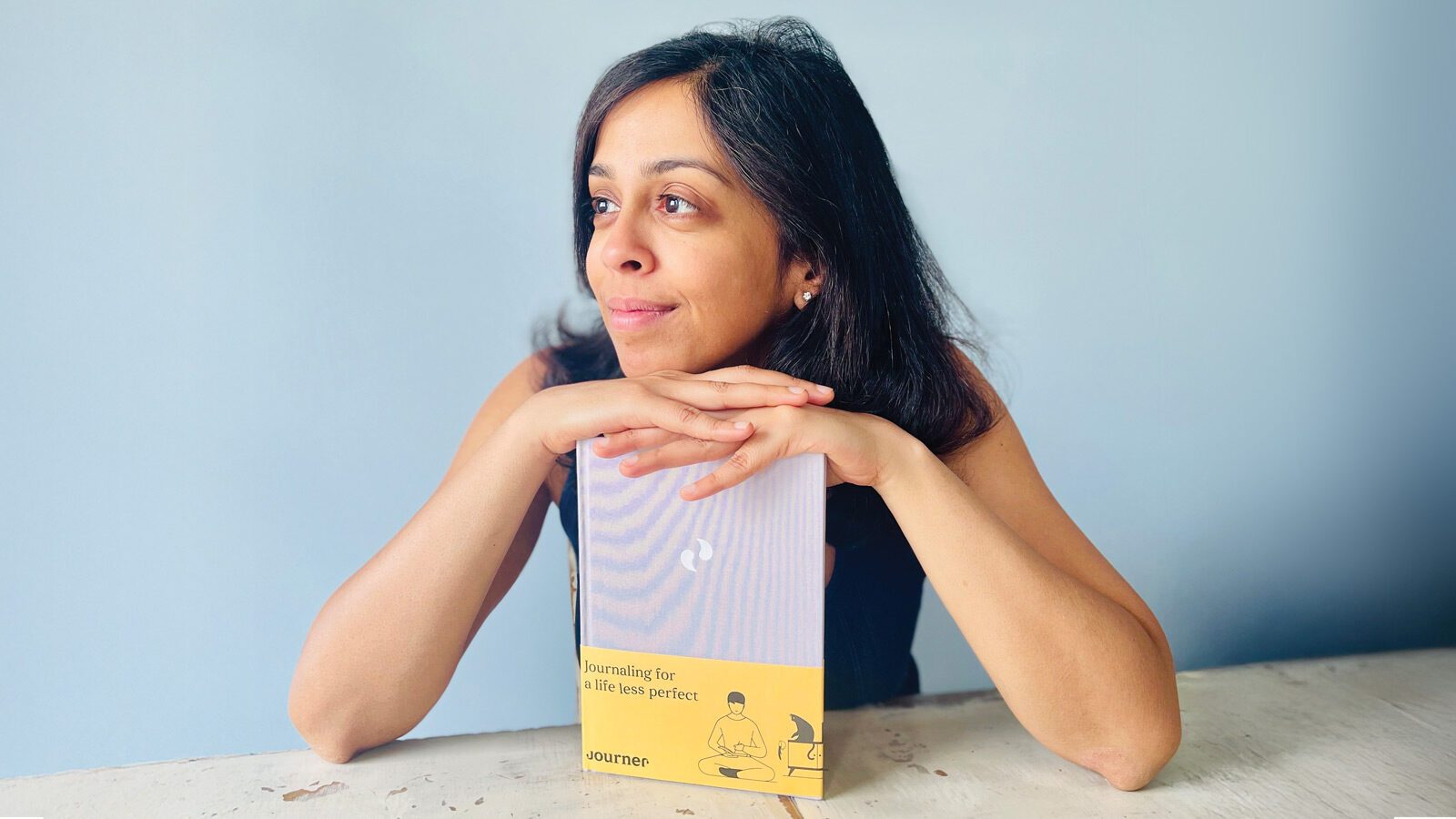 A woman sits at a table, resting her chin on her hands, with a yellow notebook titled "Journaling for a life less perfect" in front of her—ready to begin her mental journer through self-reflection.