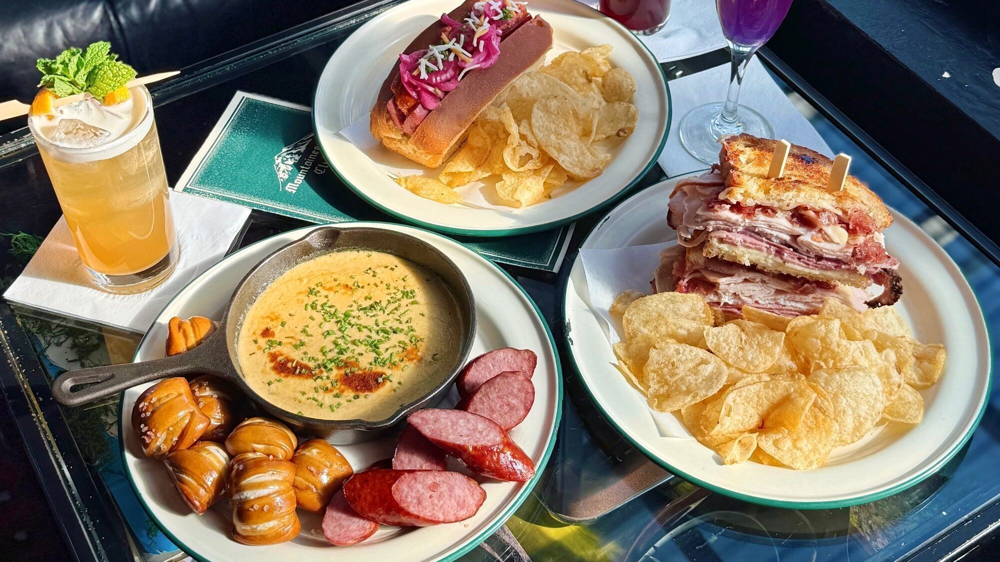 Three plates on a glass table: one with a sandwich and chips, one with sliced sausage, pretzels, and cheese dip, and one with a hot dog and chips. Two drinks are also visible.
