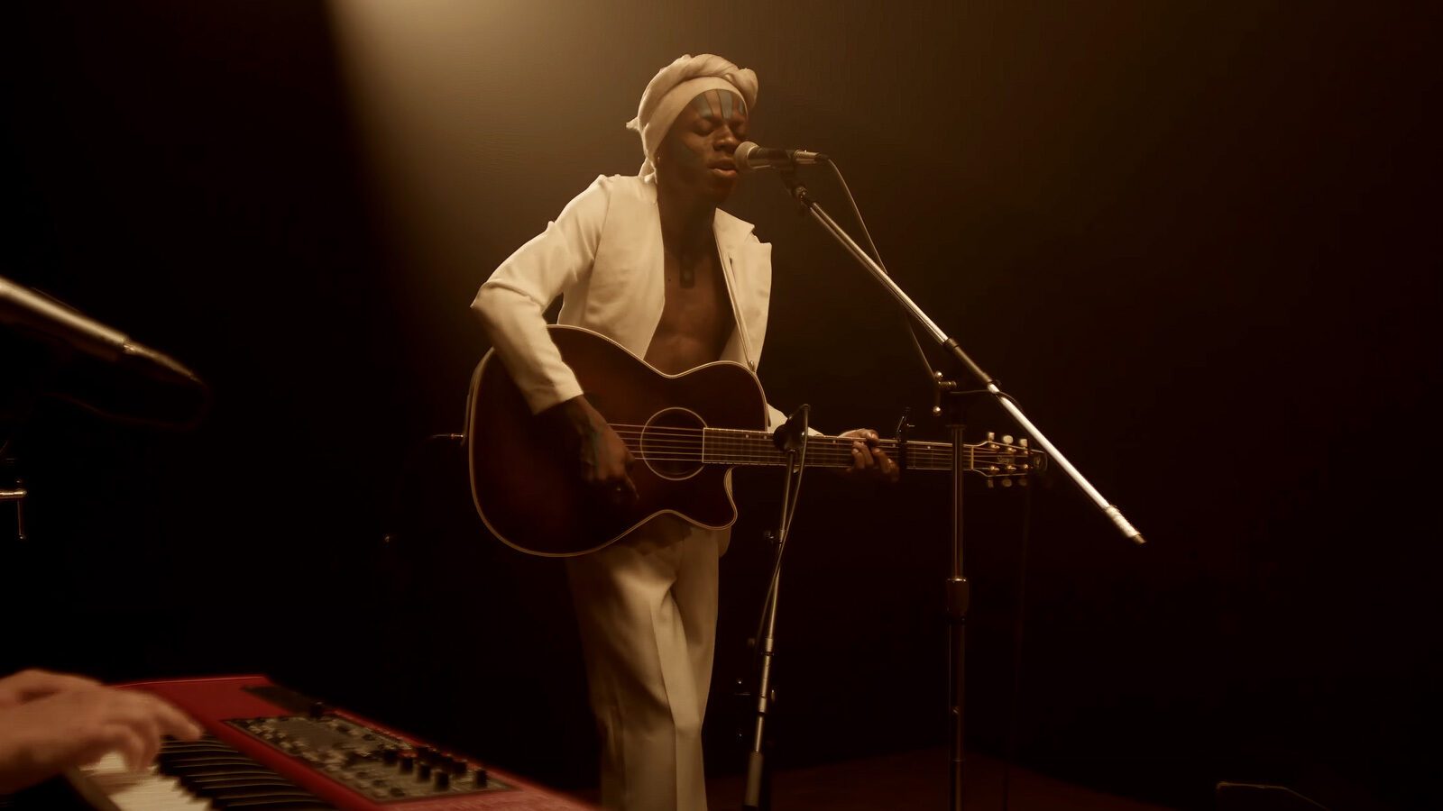 A musician in a white outfit plays an acoustic guitar and sings into a microphone on stage, delivering live music as a keyboard sits in the foreground.