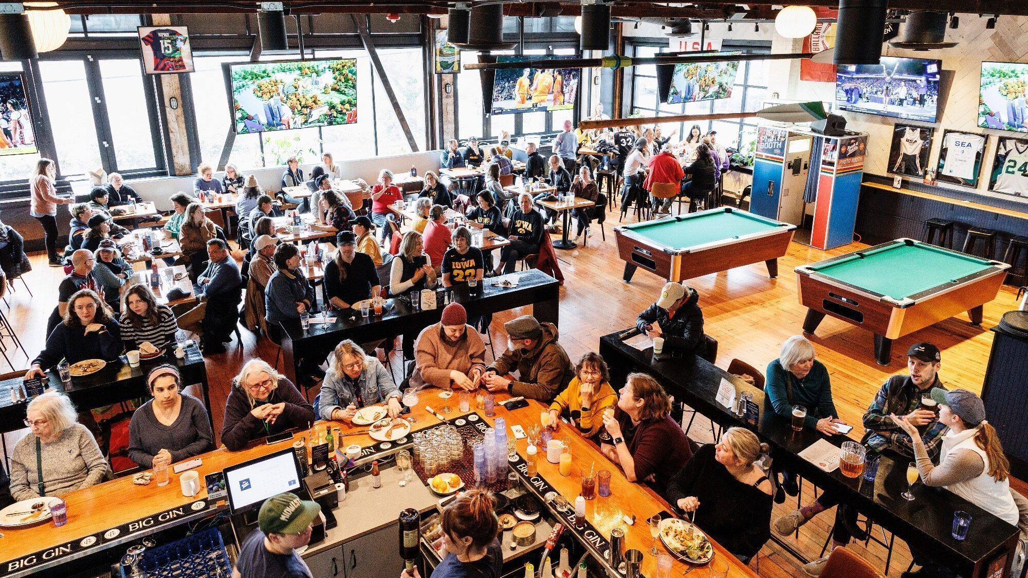 A busy sports bar with people seated at tables and the bar, eating, drinking, and watching multiple TVs; two pool tables are visible in the background.