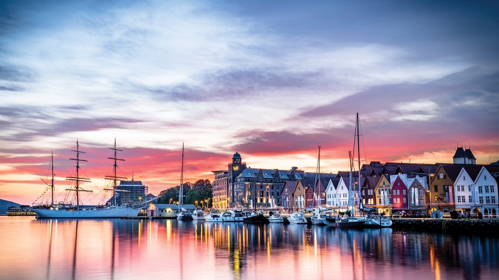 Colorful buildings and boats line a calm waterfront at sunset in Bergen, with reflections in the water and a partly cloudy sky in the background—the perfect scene for finding a home abroad.