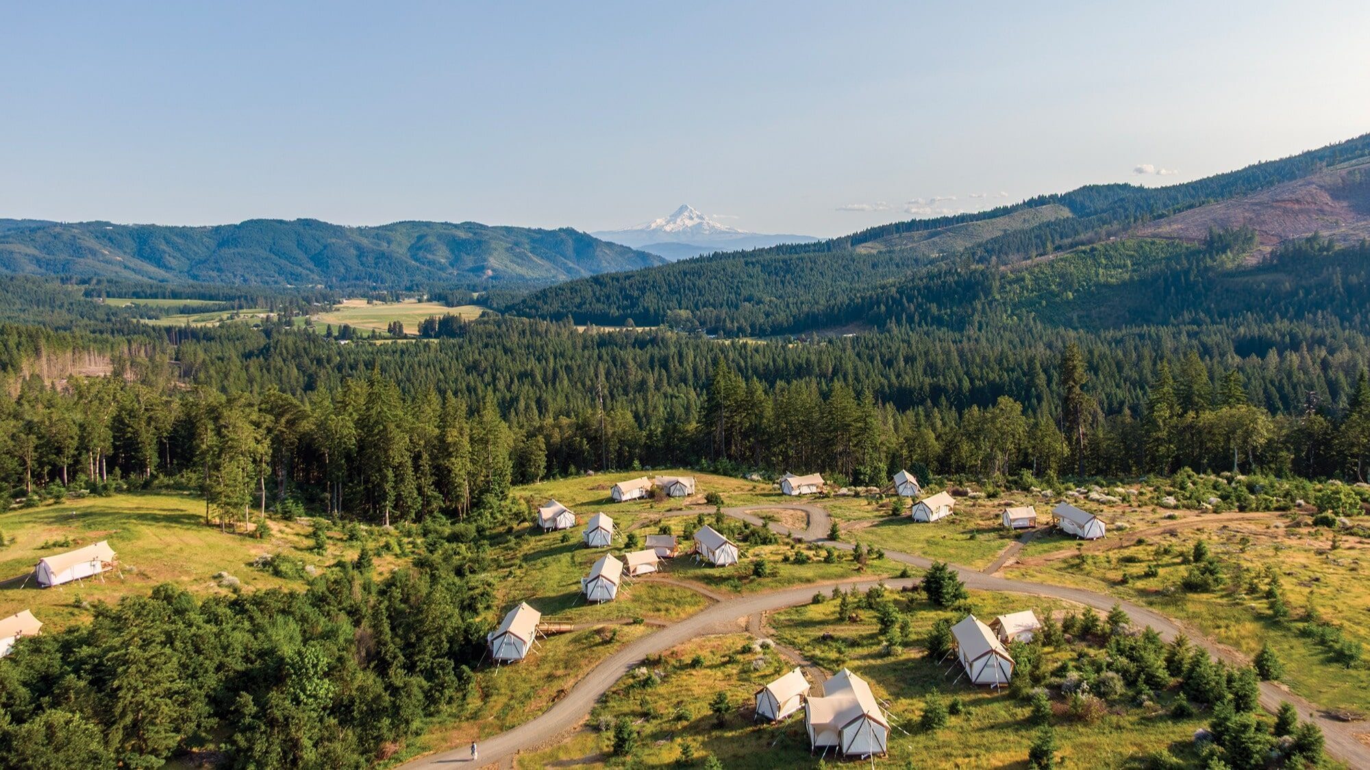 A cluster of canvas tents is set among winding paths in a grassy, wooded area, offering luxury camping with mountain views and a snow-capped peak rising in the background.
