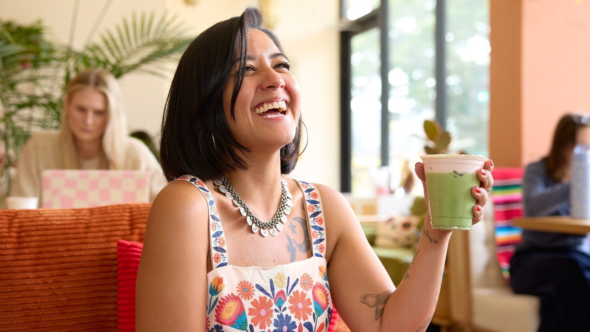 A woman in a floral dress smiles while holding a green smoothie in a brightly lit café, with other people working in the background.