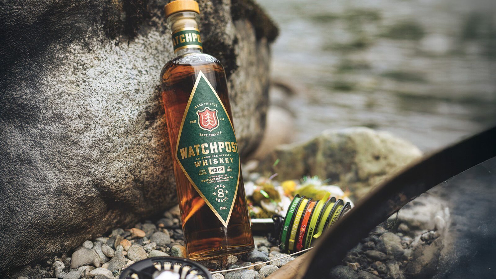 A bottle of Westland American Single Malt whiskey stands upright on rocky ground near a river, with fishing gear beside it.