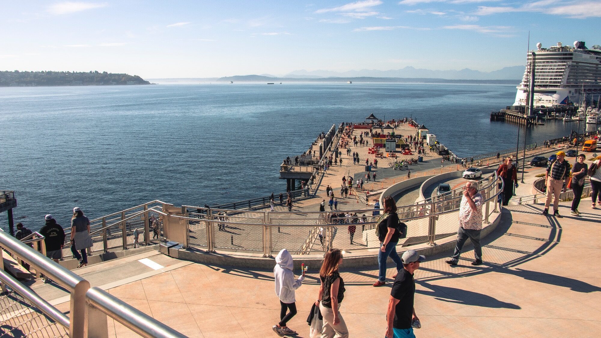 People walk along a waterfront promenade overlooking a pier extending into the water, with a cruise ship docked nearby under a clear sky.