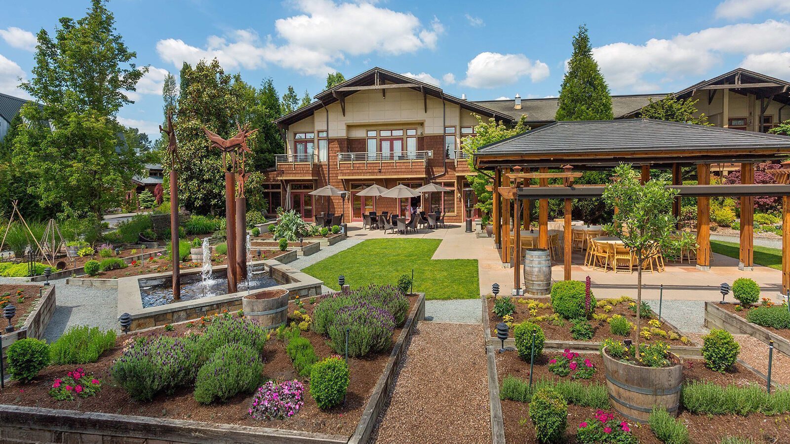 Landscaped garden with flowers and shrubs, a wooden pergola, outdoor seating for relaxation, and a large building in the background under a partly cloudy sky—perfect for enjoying Woodinville’s wine country ambiance.