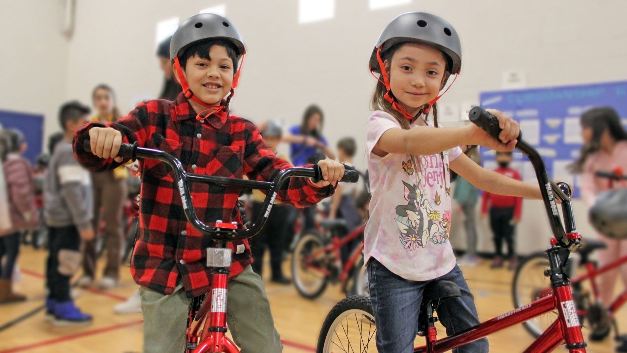 Two children wearing helmets sit on red bicycles in a gymnasium, surrounded by other people in the background.