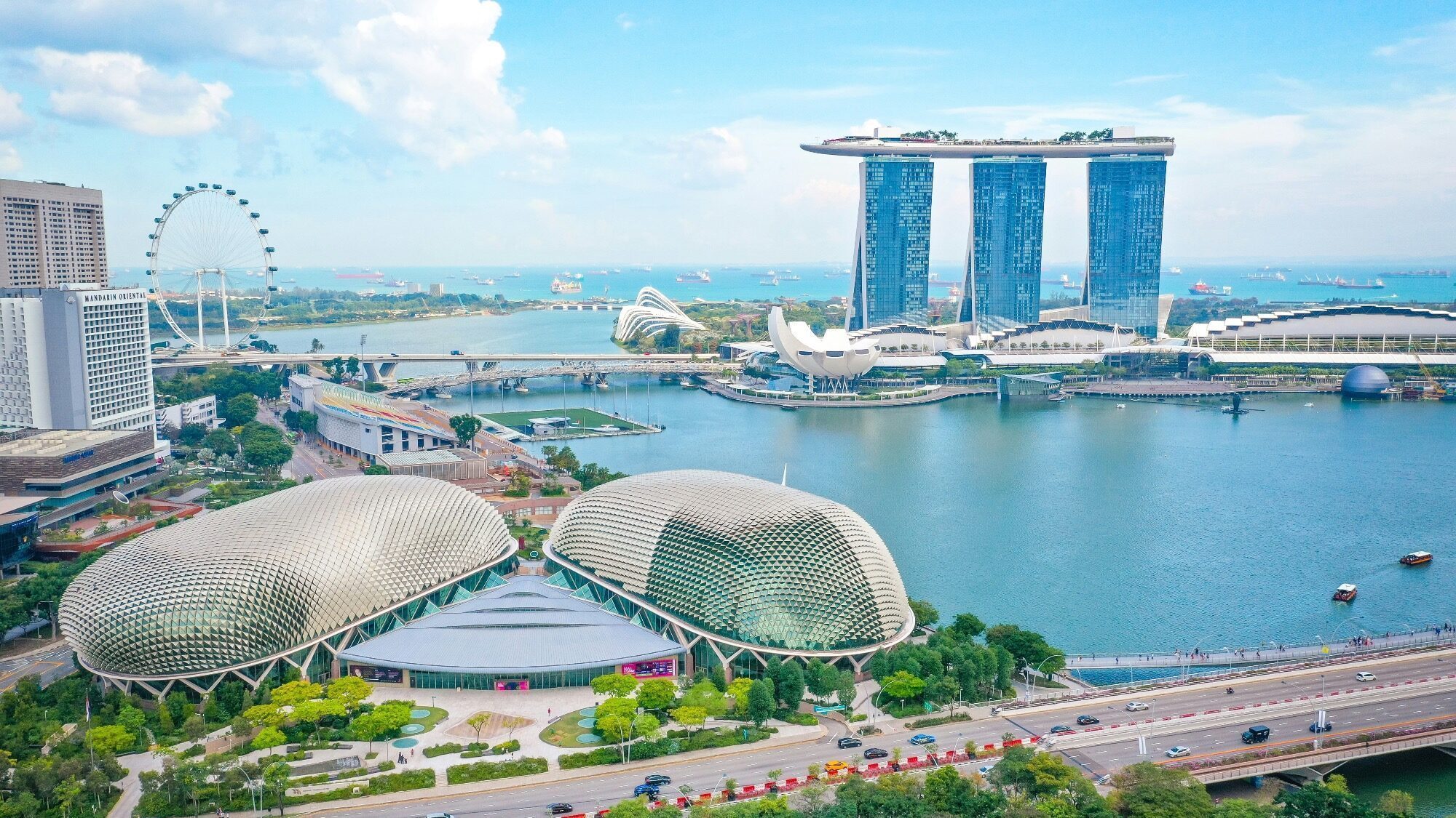 Aerial view of Singapore Marina Bay with the Esplanade Theatres, Marina Bay Sands hotel, Singapore Flyer Ferris wheel, and cityscape under a blue sky.