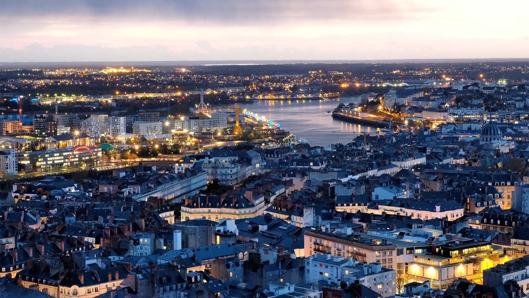 A cityscape at dusk with illuminated buildings, winding streets, and a river running through the center, reflecting city lights under a cloudy sky.