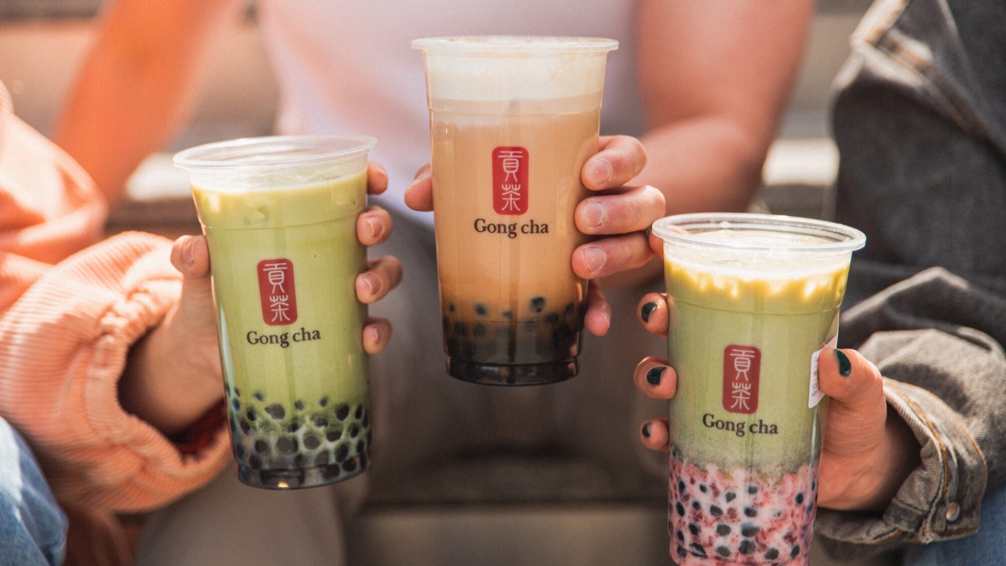 Three people holding different Gong Cha bubble tea drinks, each with tapioca pearls, outdoors.