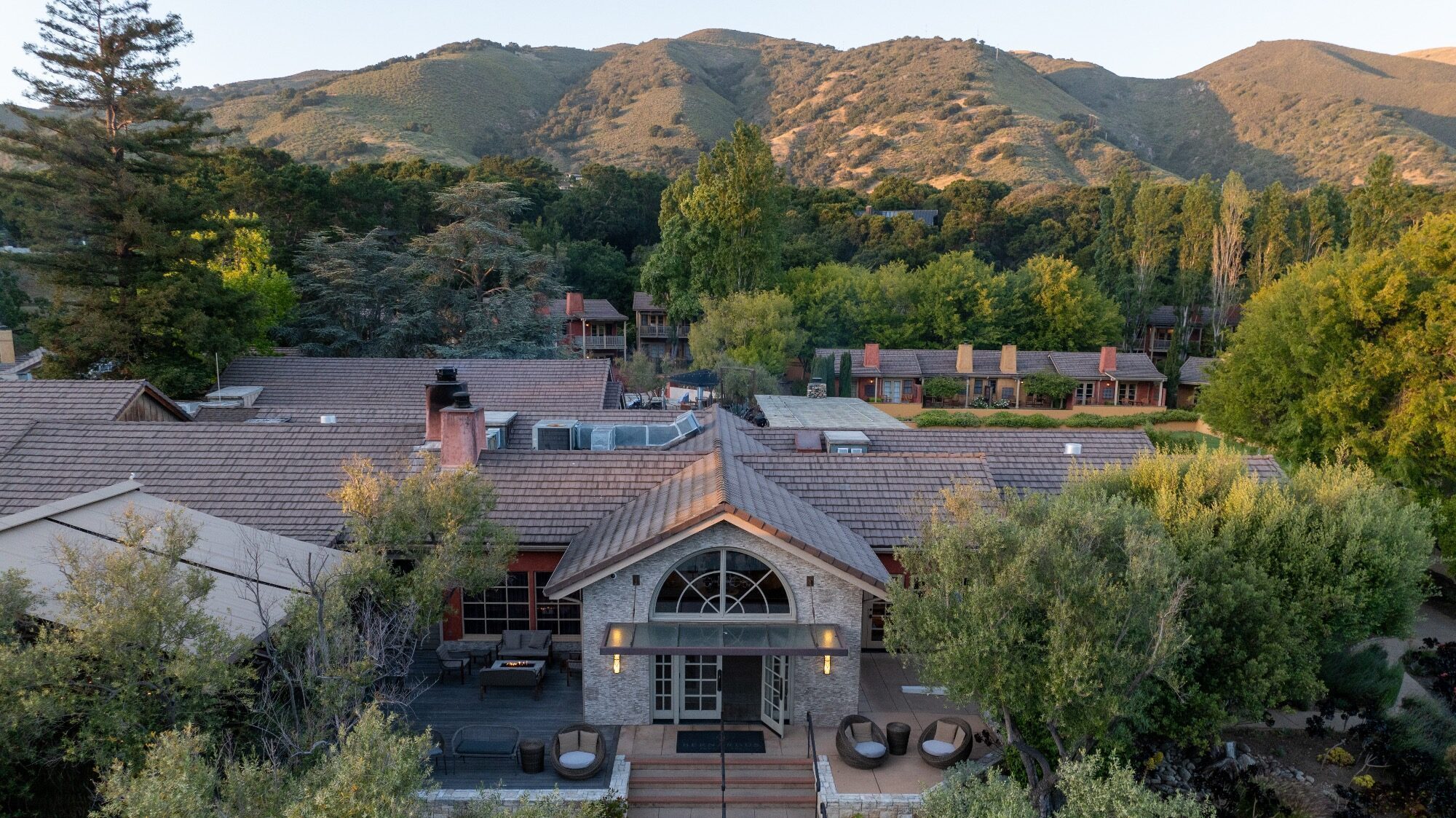 A large house with a tiled roof sits among trees, with additional buildings and green hills visible in the background.