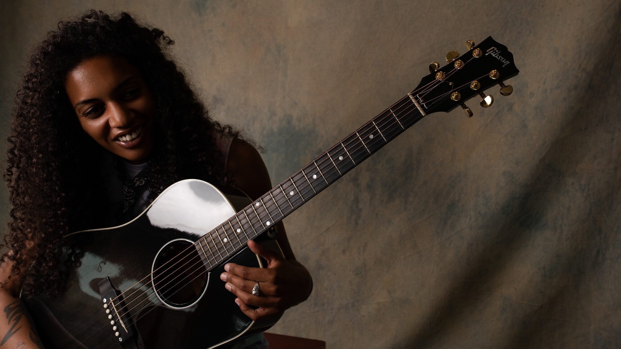 A person with long curly hair smiles while holding and playing a black acoustic guitar against a neutral background.