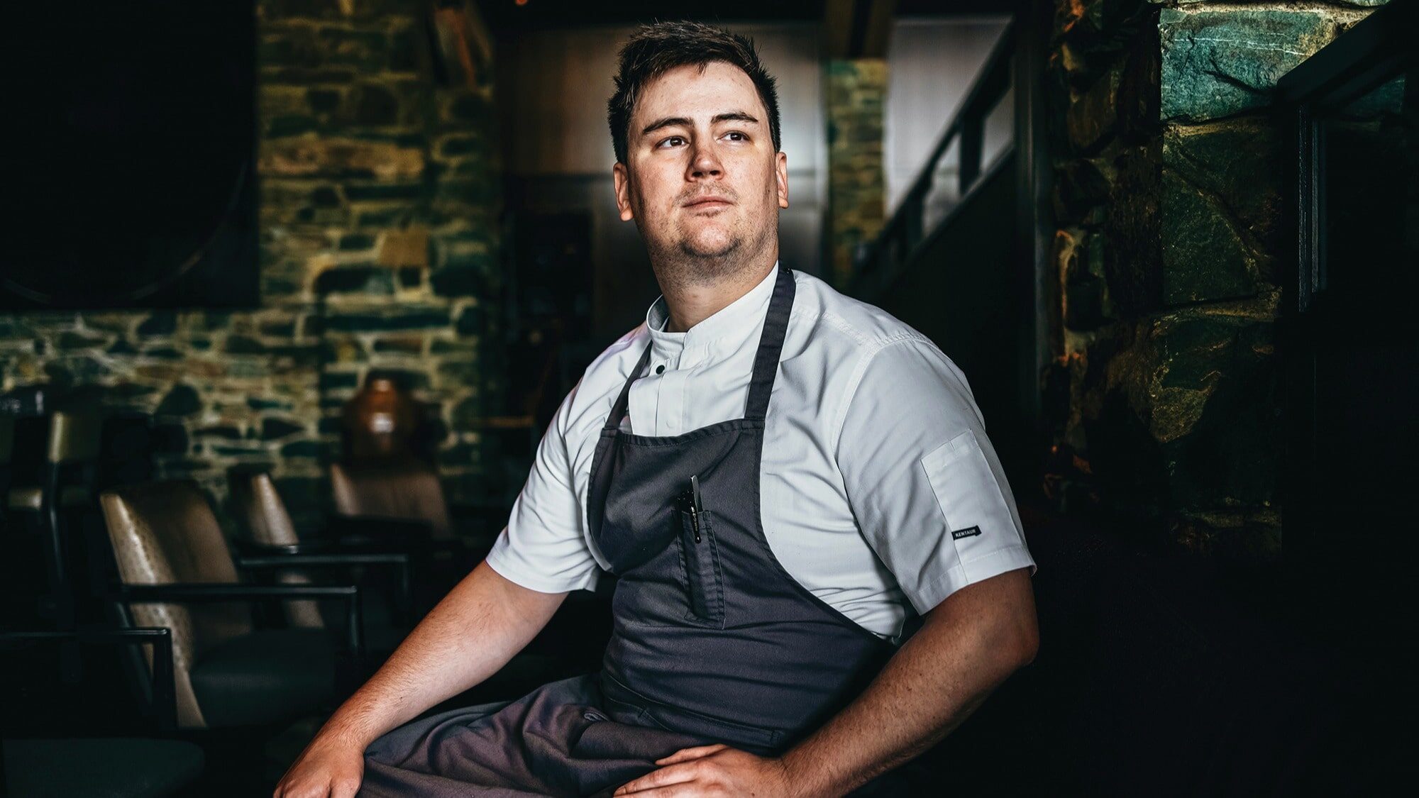 A chef in uniform sits on a chair in a rustic restaurant with stone walls, embodying the spirit of Hometown Harvest, looking off to the side with a neutral expression.