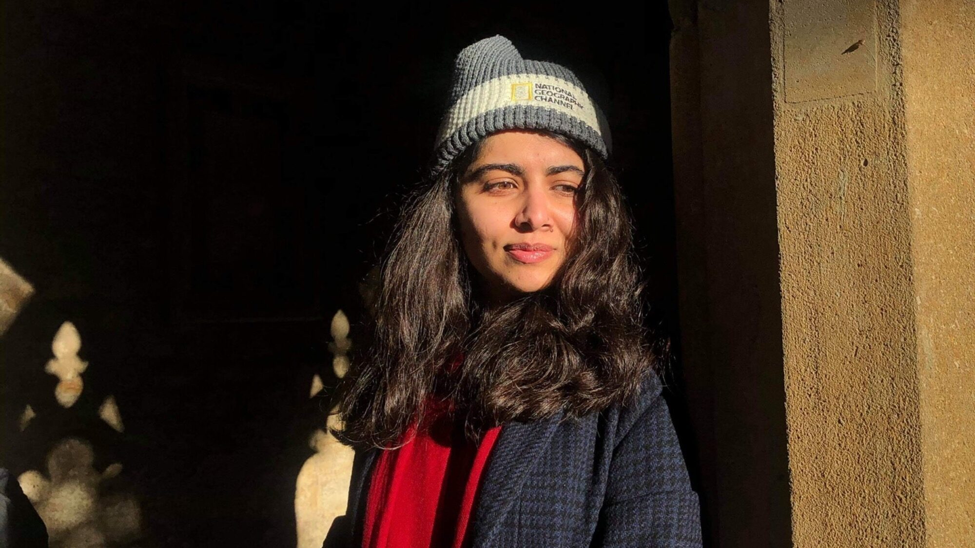 A woman wearing a gray beanie and dark coat stands in sunlight beside a stone wall, looking to the side.