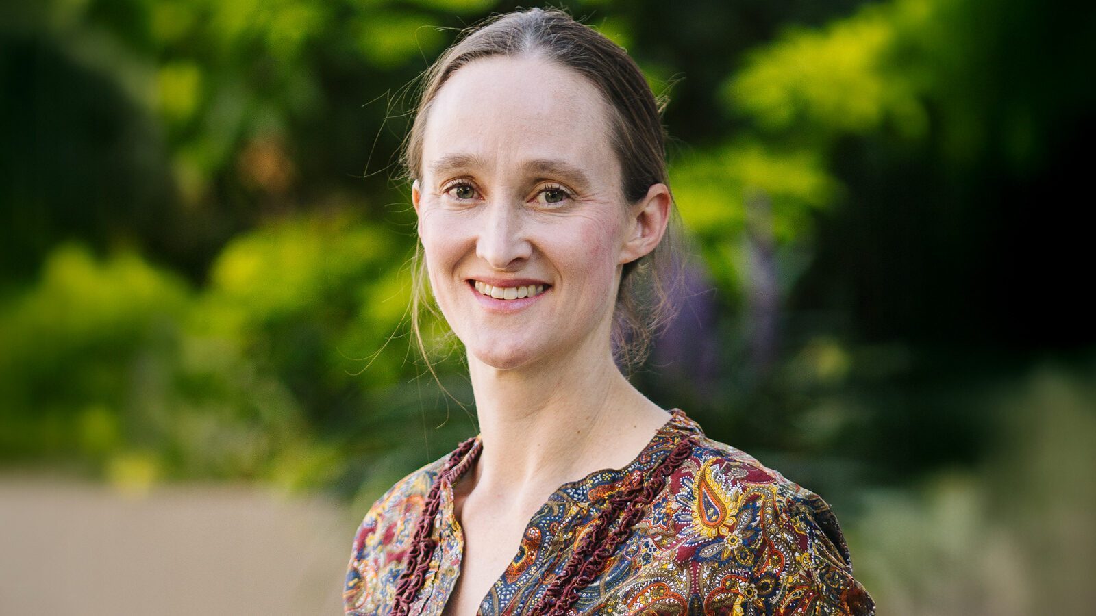 Katie Wilson, a woman with light skin and brown hair tied back, is smiling outdoors in a patterned blouse, with greenery blurred in the background.