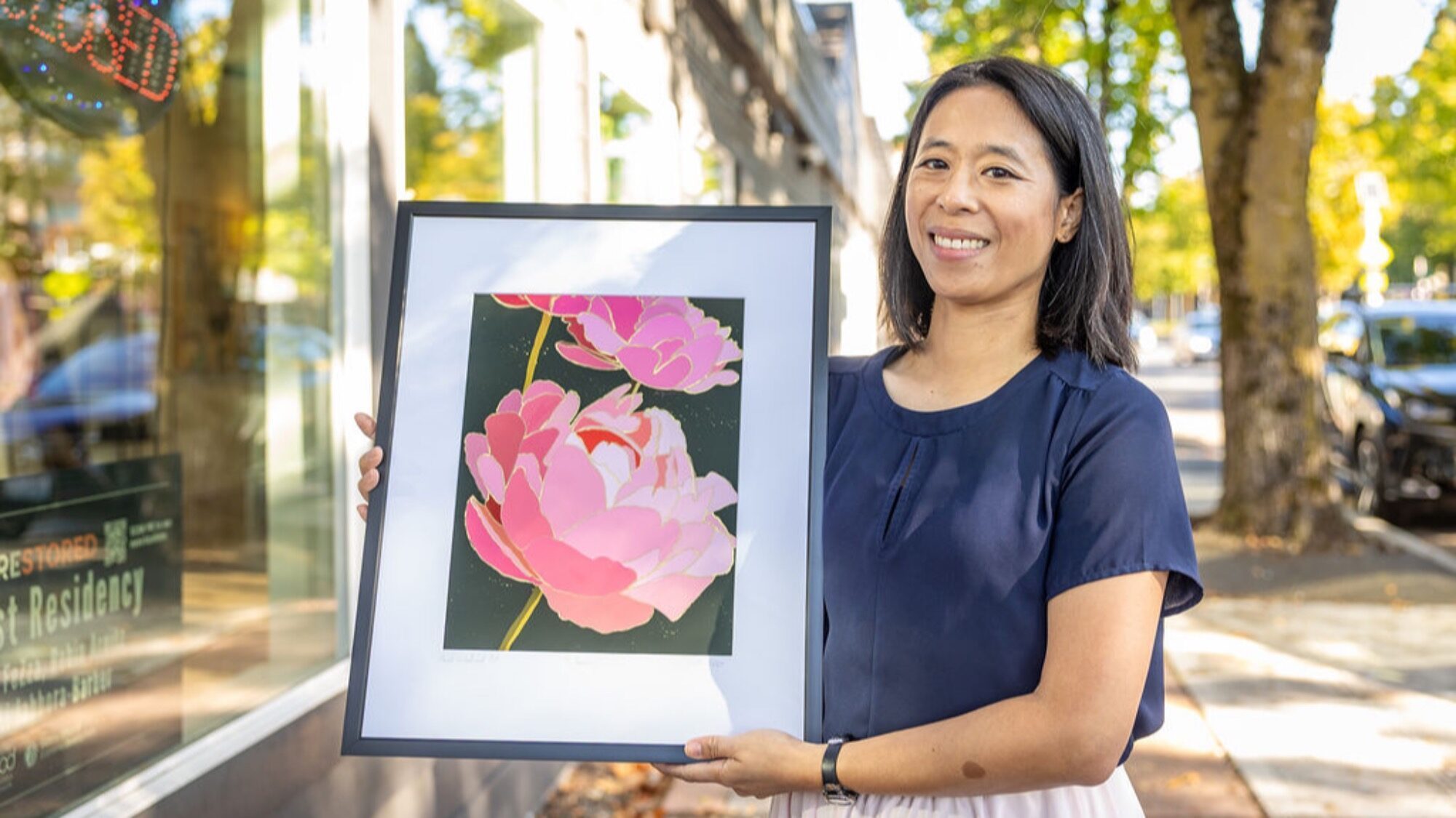 A woman stands outdoors holding a framed artwork featuring large pink flowers against a dark background.