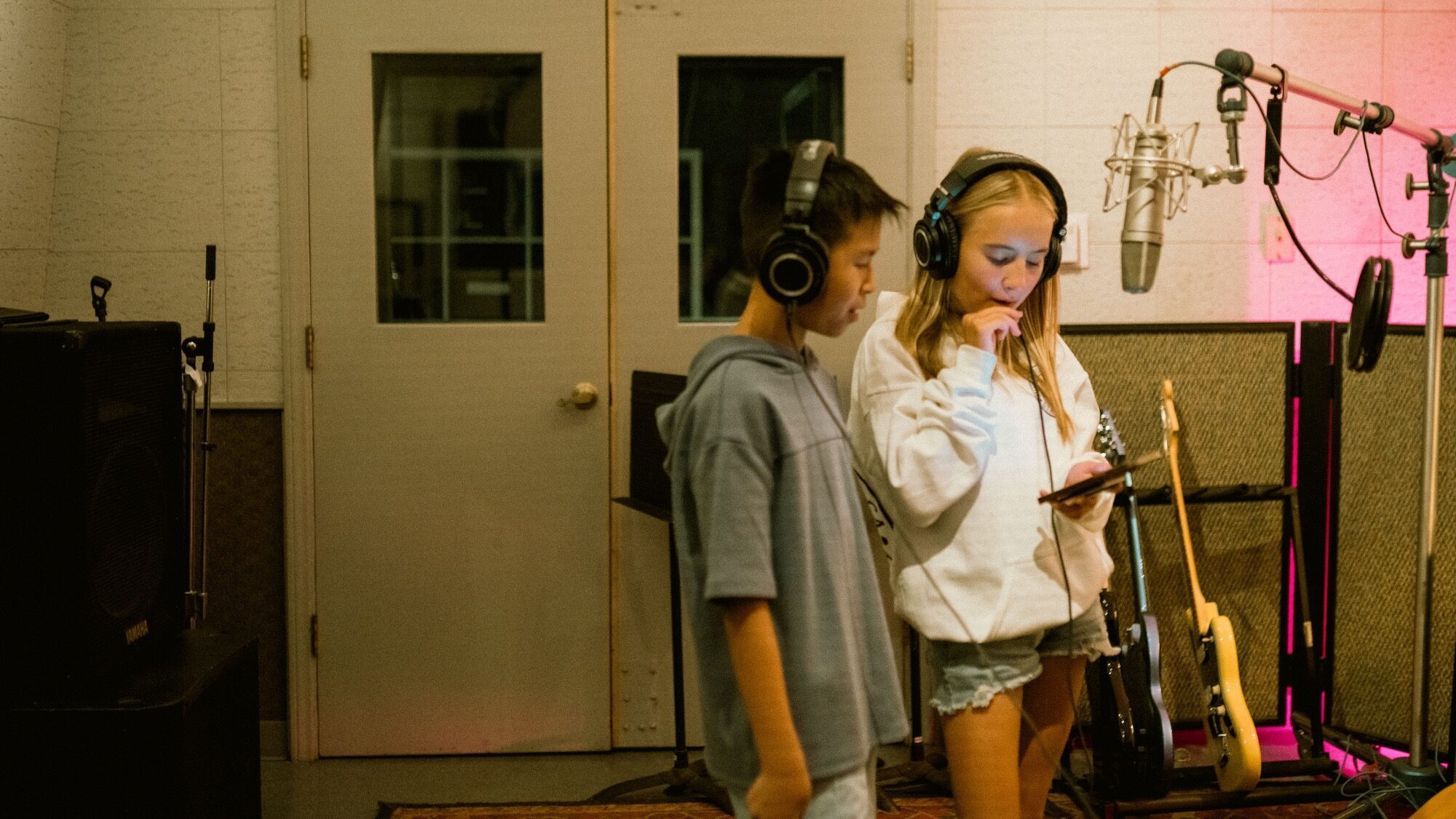Two children wearing headphones stand in a recording studio next to microphones and guitars; one looks at a phone while the other looks on.