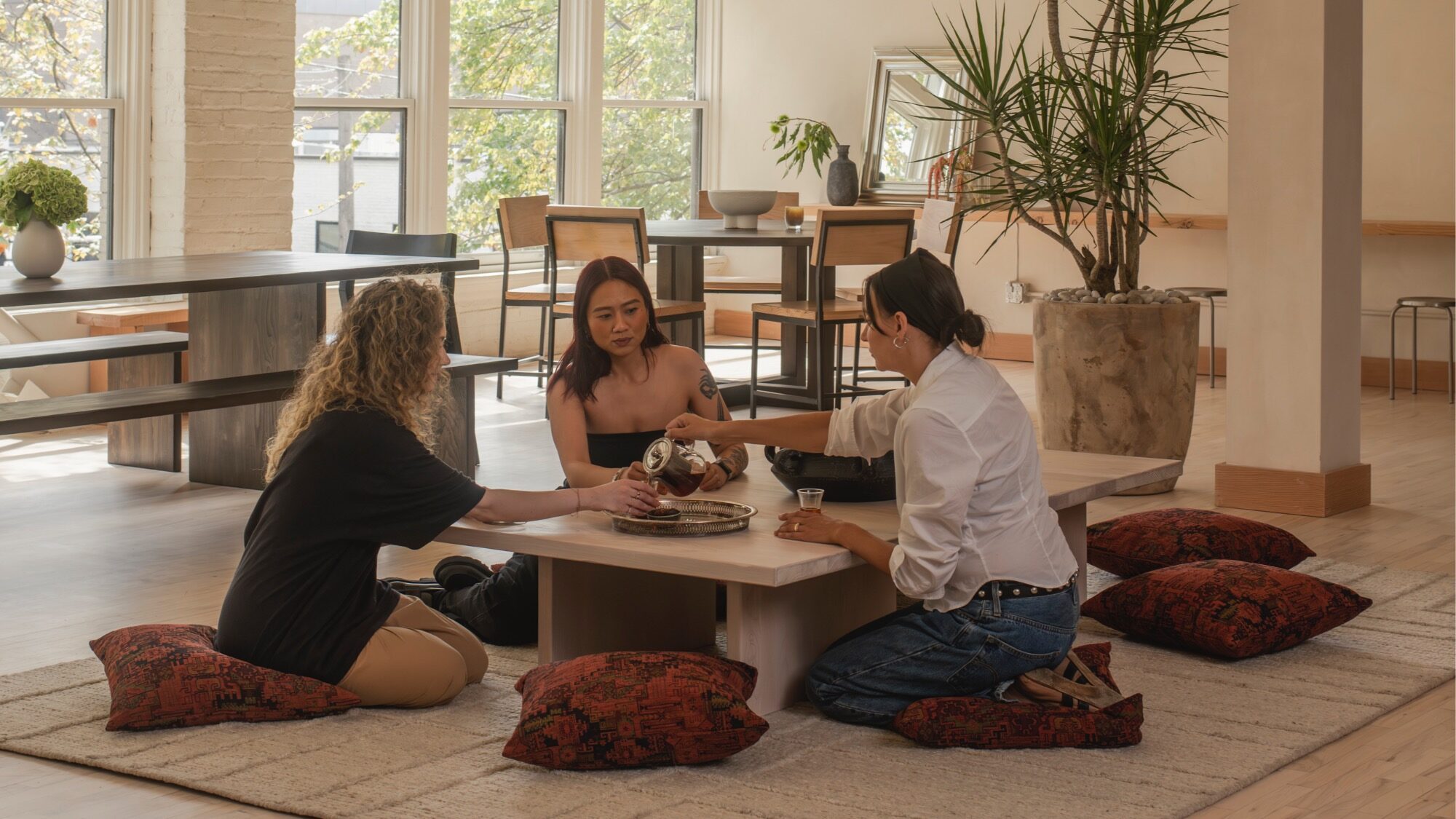 Three women sit on floor cushions around a low table, pouring tea in a sunlit room with large windows, modern furniture, and potted plants.