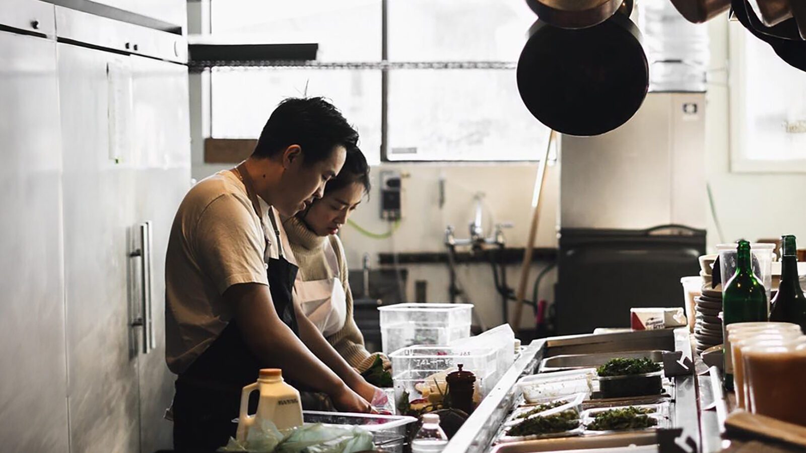 Two people are preparing food at a kitchen counter, surrounded by various cooking supplies and ingredients.
