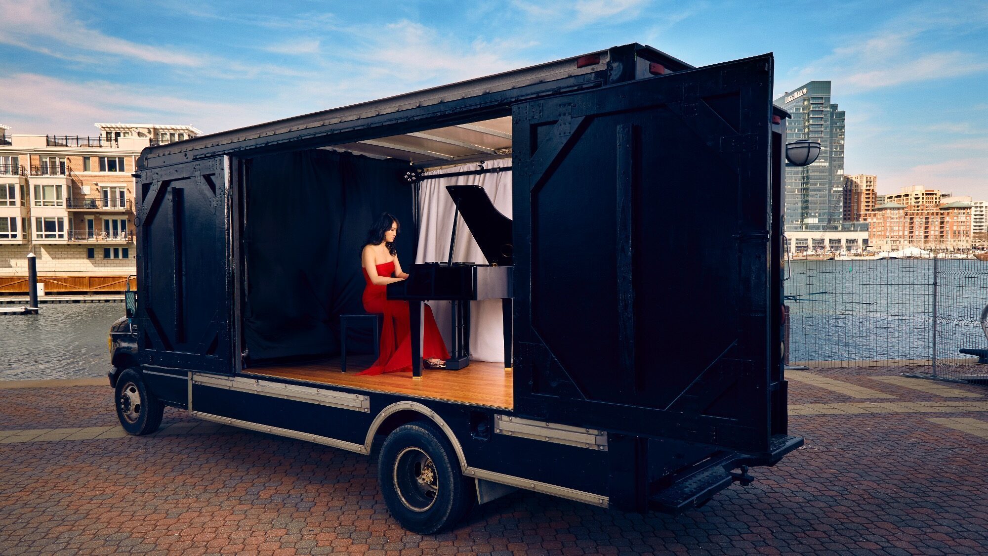 A woman in a red dress plays a grand piano inside a modified truck with open sides, parked by a waterfront with buildings in the background.