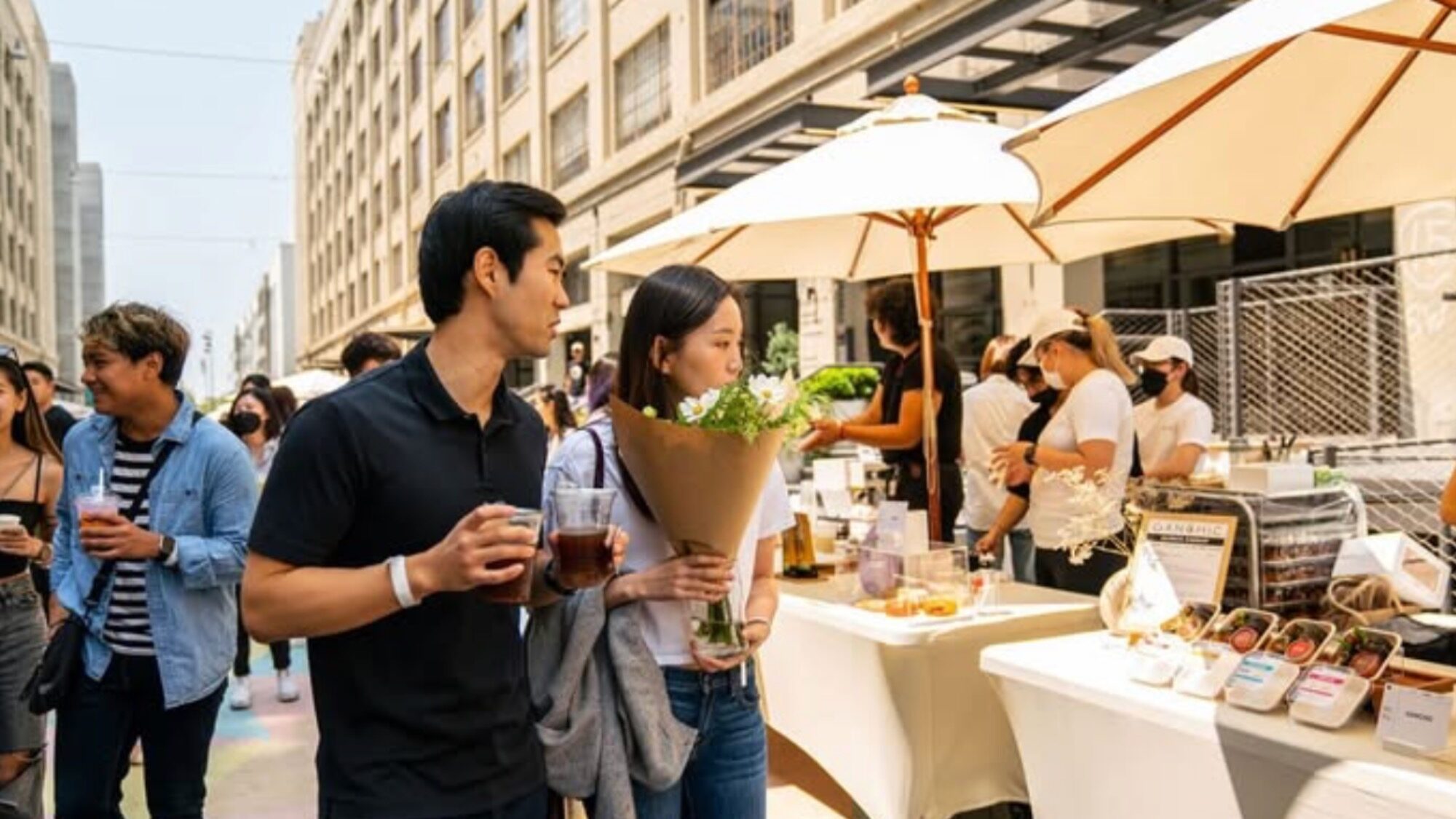 A man and woman walk past outdoor market stalls at Seattle's MAUM Market; the woman holds a bouquet of flowers and both hold drinks. Other people browse the stalls under umbrellas.