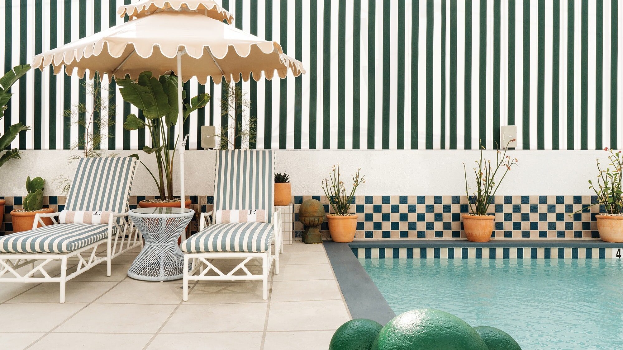 Two striped lounge chairs and a scalloped umbrella sit by a pool in Los Angeles, with potted plants and a green-and-white striped wall in the background.