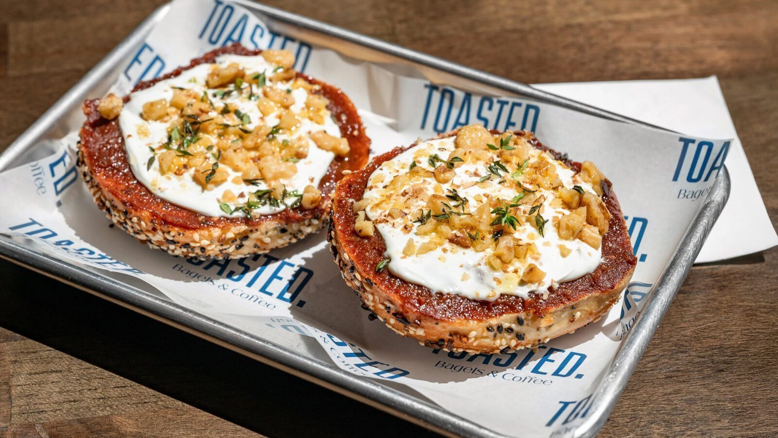 Two everything bagel halves topped with a thick layer of whipped cream cheese, chopped nuts, and herbs, served on a metal tray lined with branded paper—a must-try for Halal Bagels fans at Restaurant Roundup.