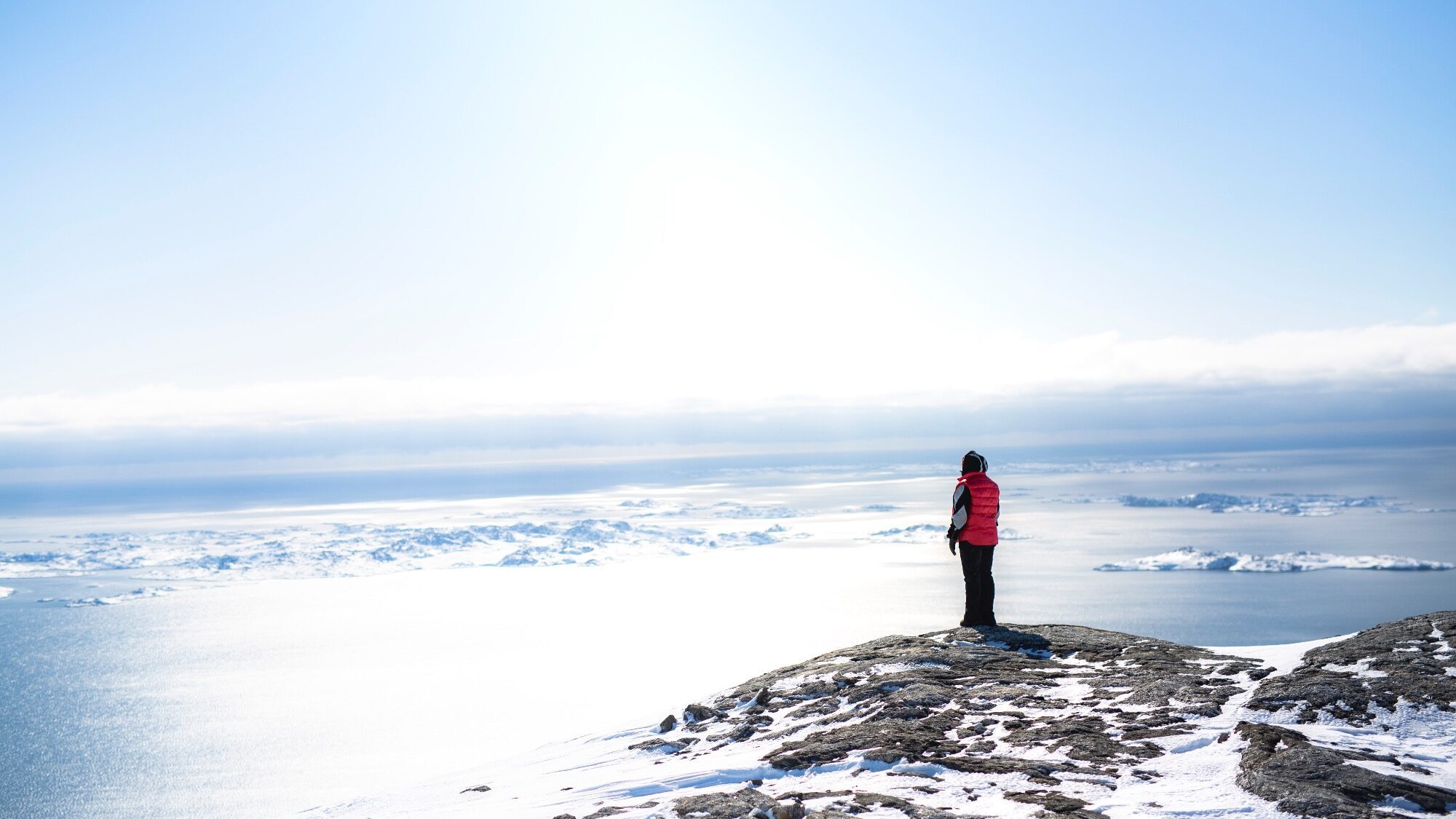 A person in a red jacket stands on a snowy cliff overlooking a vast, icy landscape and ocean under a bright sky.