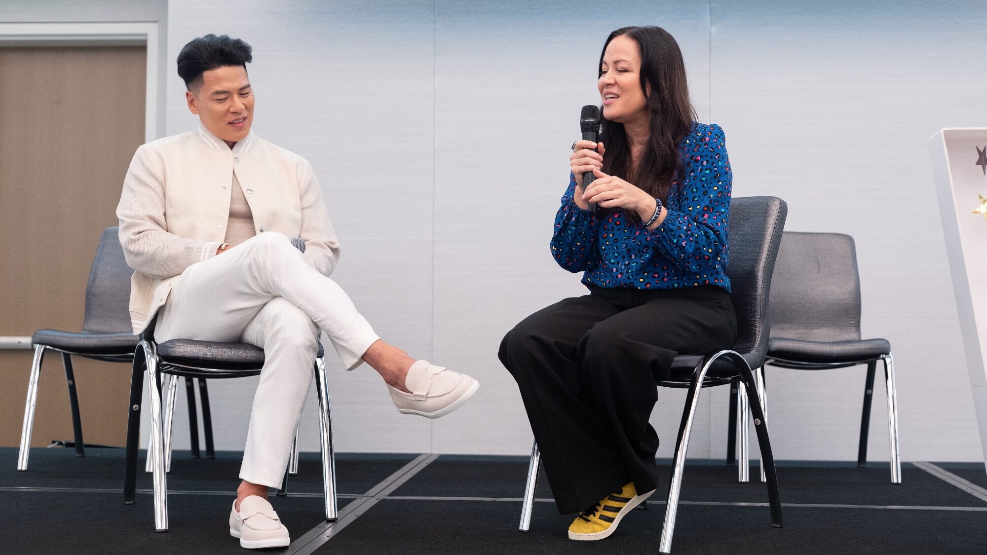 A man and a woman sit on chairs on a stage; the woman holds a microphone and speaks while the man listens attentively.