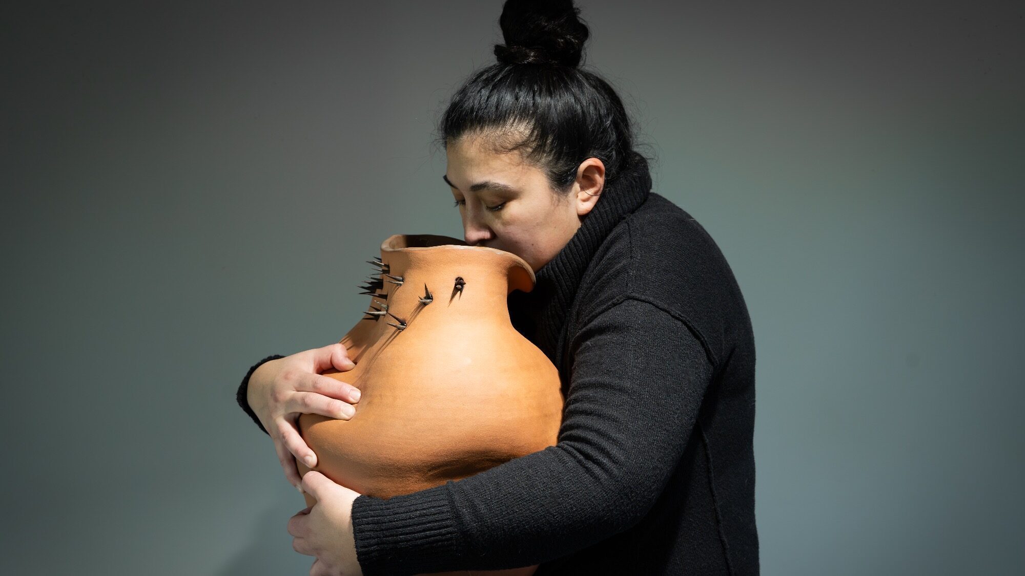 Person in a black turtleneck holds and leans face against a large, irregularly shaped clay pot with cracks and small black objects attached.