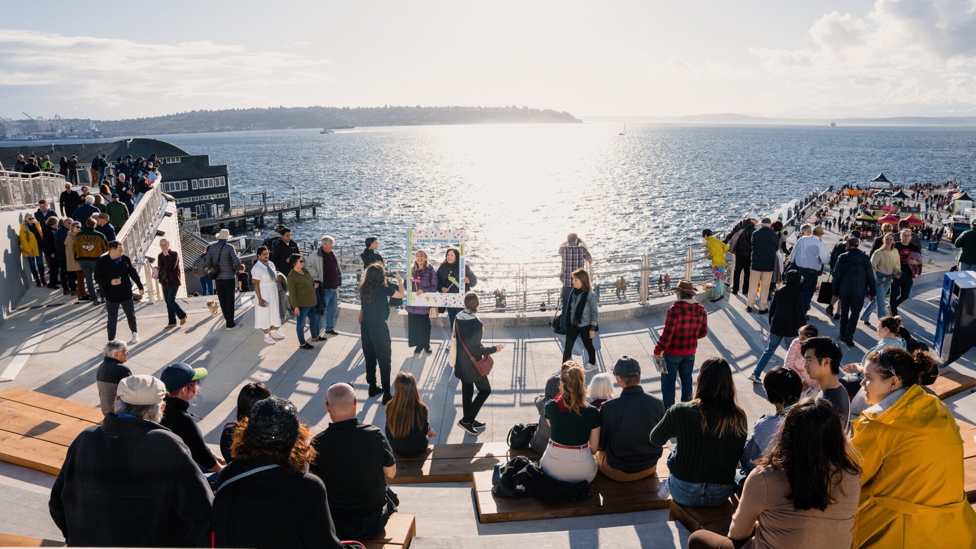 A crowd of people gathers on tiered seating and walkways by the waterfront under a bright sky, with sunlight reflecting off the water.