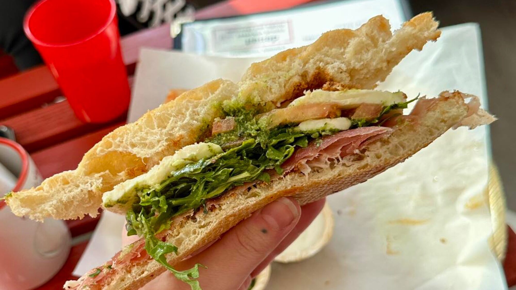 A hand holds an Italian Sandwich with arugula, mozzarella, tomato, prosciutto, and pesto on ciabatta bread—perfect for a light lunch at your favorite restaurant.