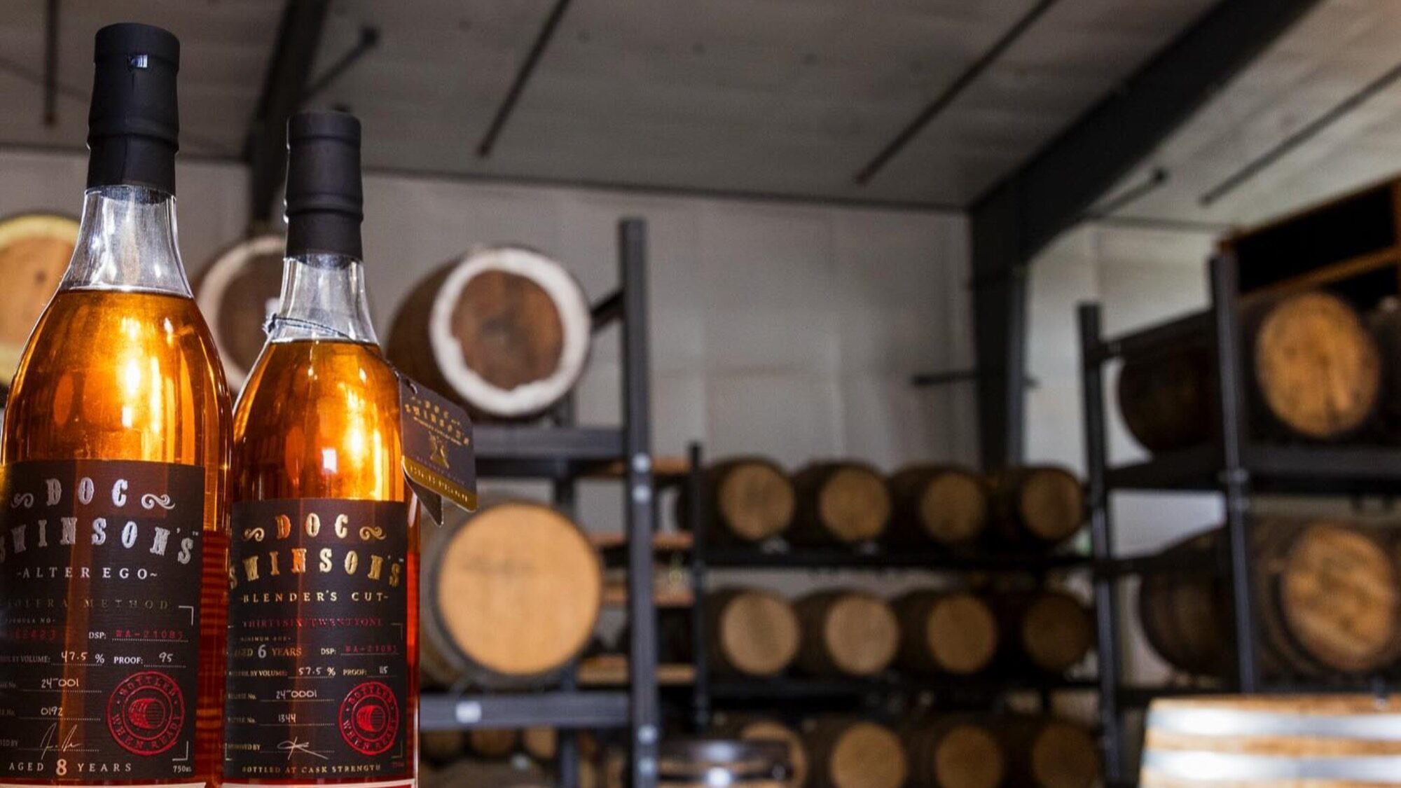 Two bottles of whiskey labeled "Doc Swinson's" are displayed in front of shelves stacked with aging barrels in a distillery storage room.