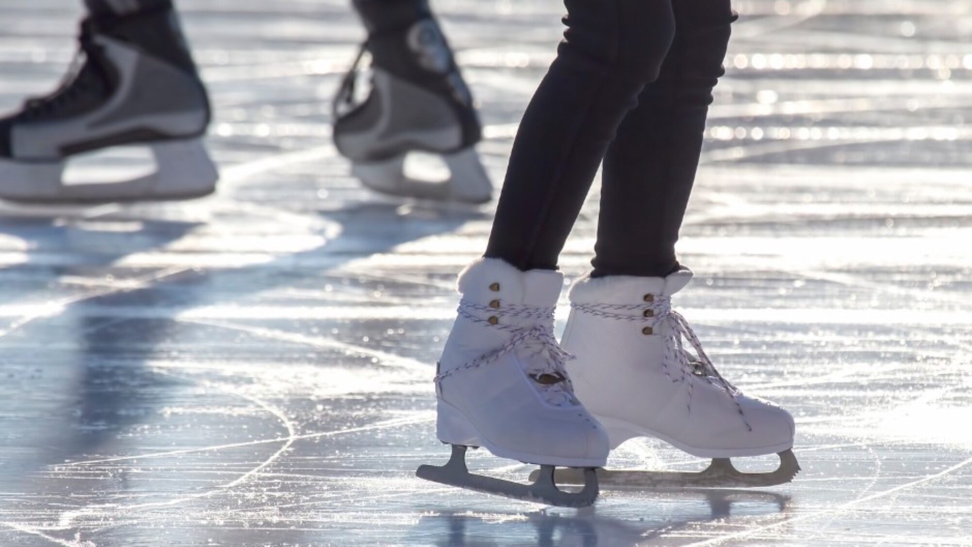 Close-up of people ice skating outdoors, showing their legs and ice skates on a shiny, sunlit ice surface.