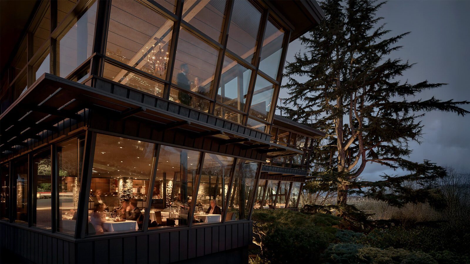 Modern glass-walled Seattle restaurant at dusk with diners inside, surrounded by large trees and outdoor greenery, and warm interior lighting glowing through the windows—a perfect setting for a special occasion restaurant like Canlis.